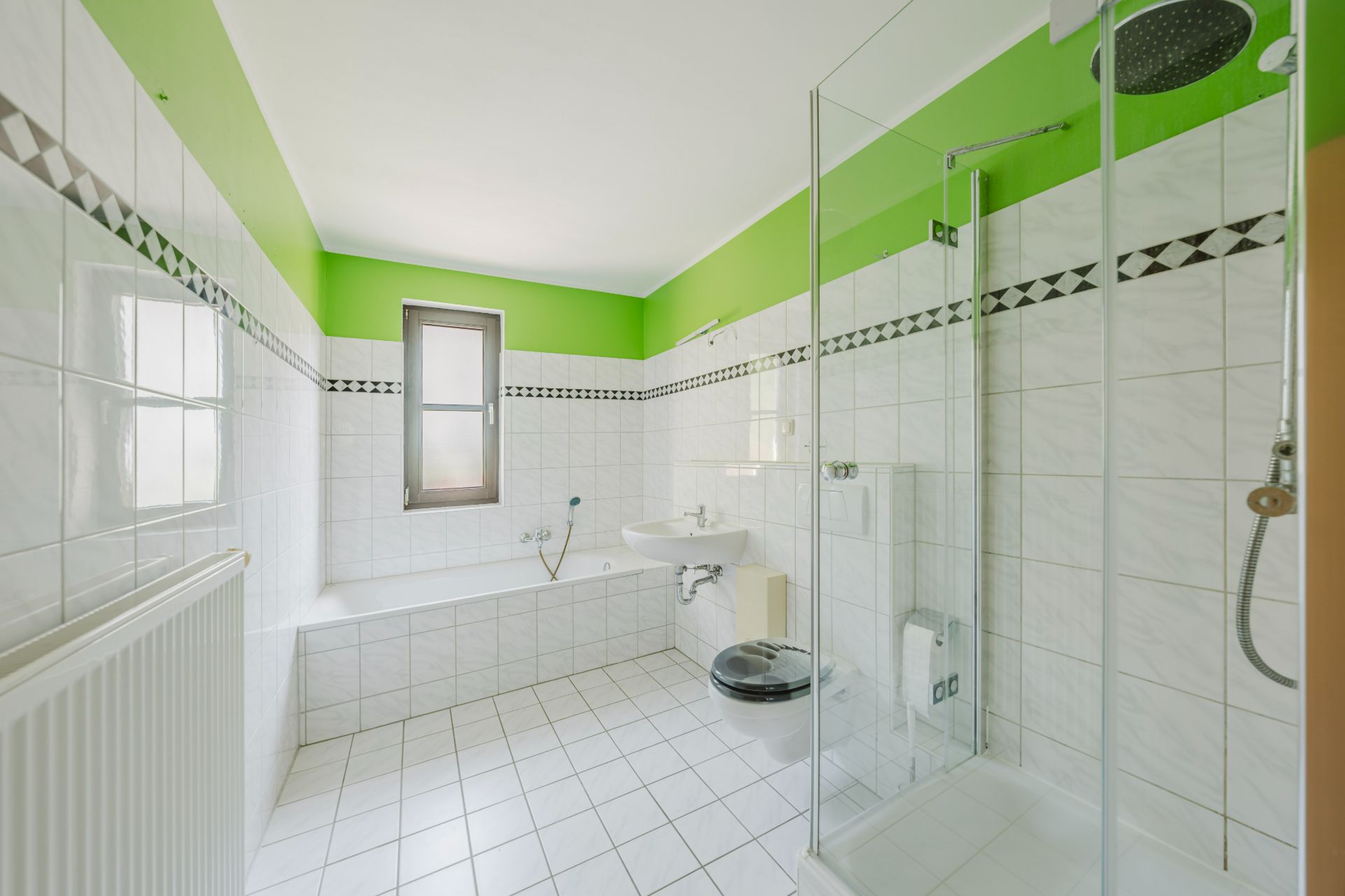 Helles Badezimmer mit Badewanne, Dusche, WC, Waschbecken und Fenster, grüne Wandfarbe.