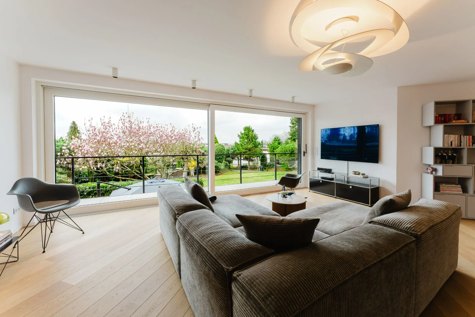 Helles Wohnzimmer mit großem Sofa, Fernseher und Blick auf einen Garten durch Panoramafenster.