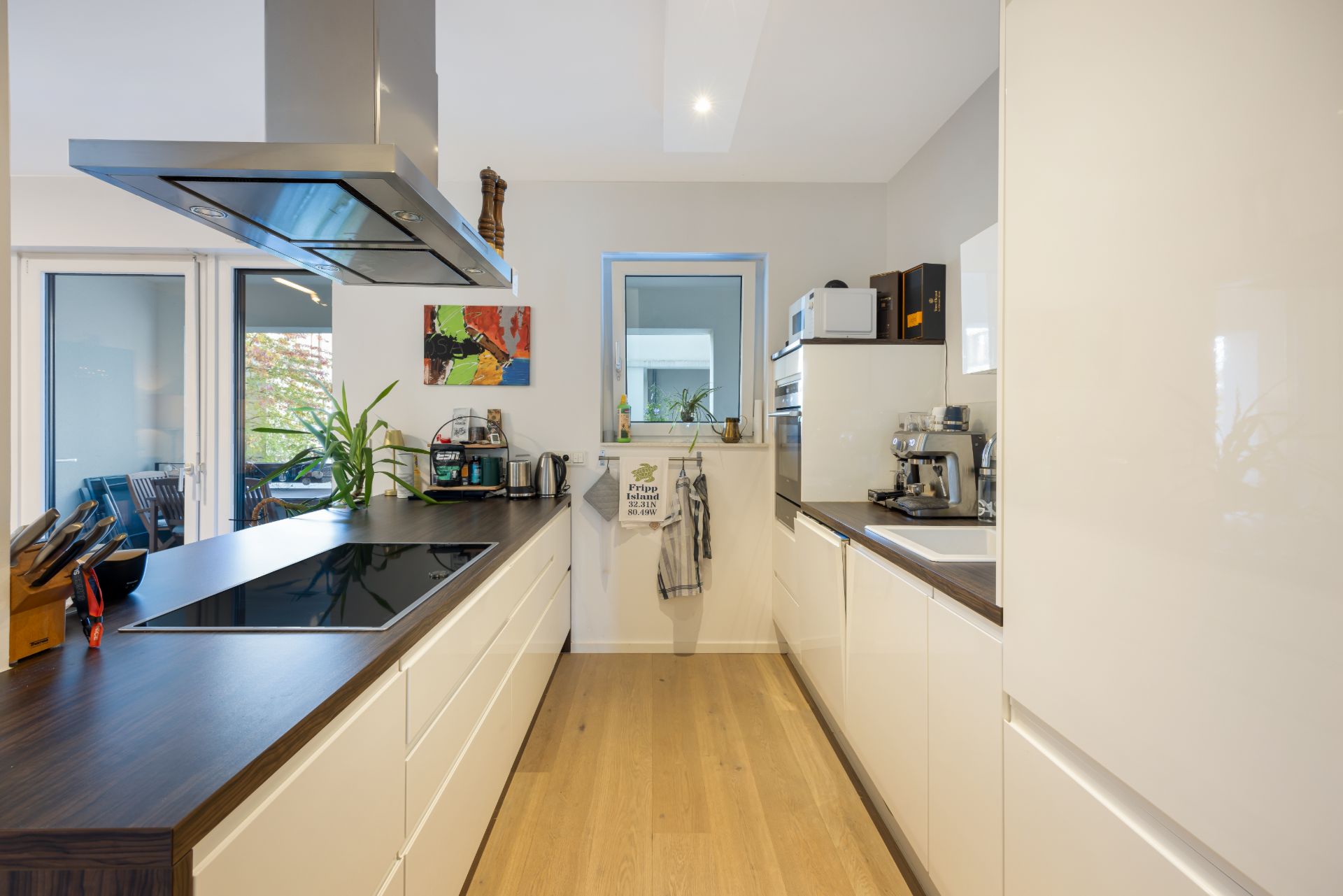 Moderne Küche mit weißen Schränken, Holzboden und Fenstern, Kaffeemaschine und Kochfeld sichtbar.