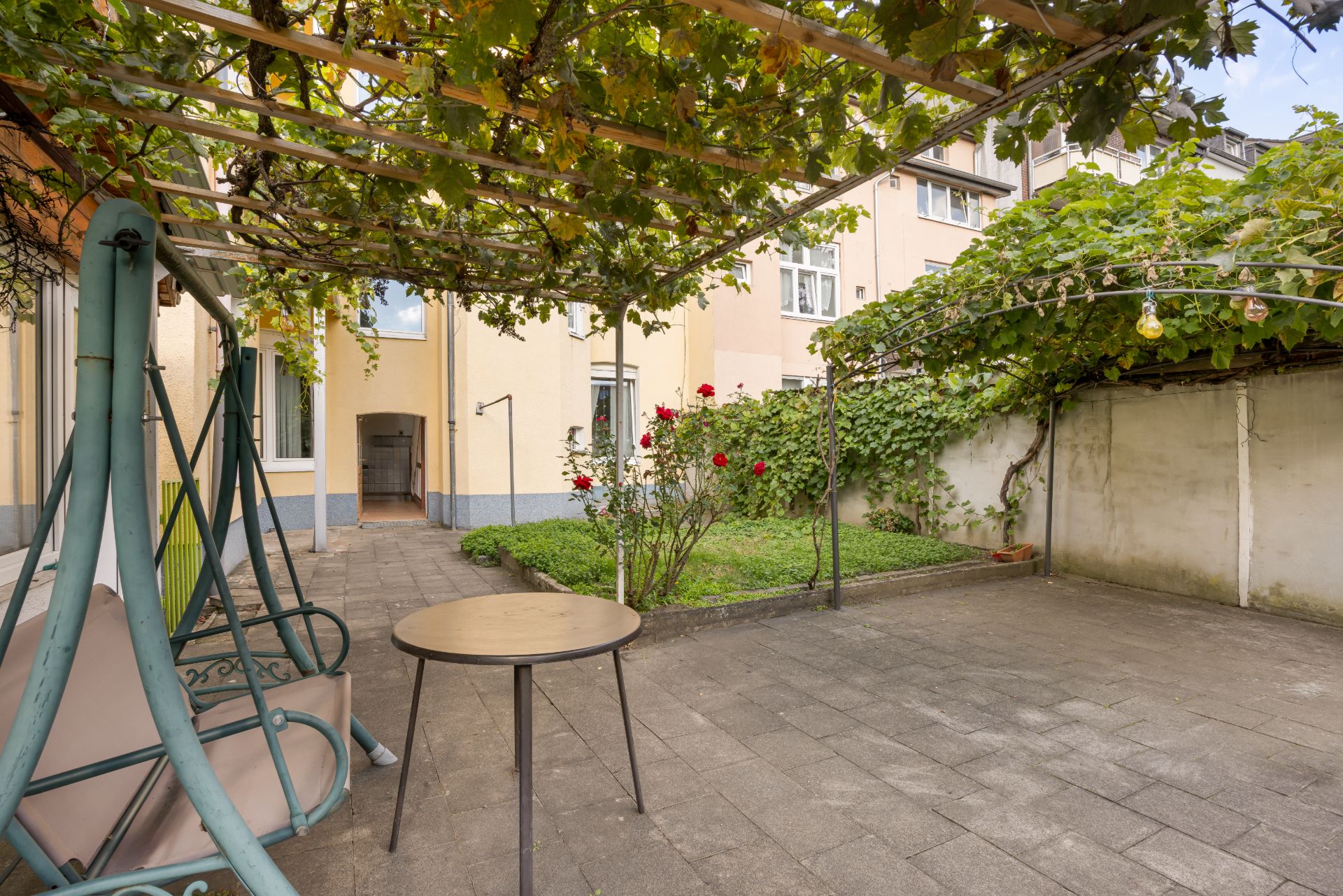 Gepflasterter Innenhof mit Hollywoodschaukel, Tisch, Rosenstrauch und Rankgitter mit Pflanzen.