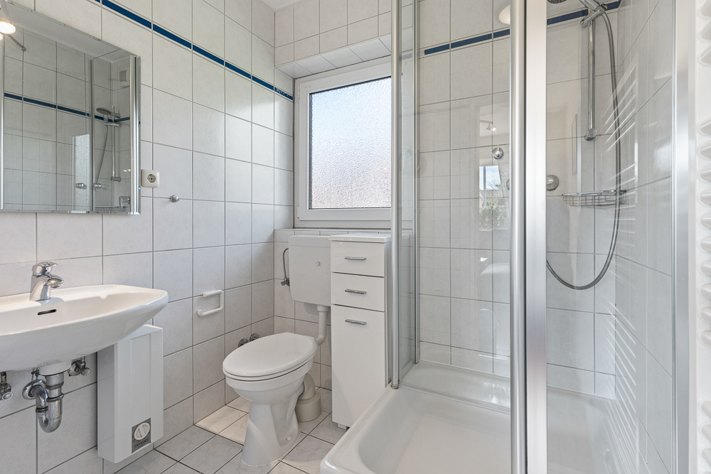 Modernes, weiß gefliestes Badezimmer mit Dusche, WC, Waschbecken und Fenster.