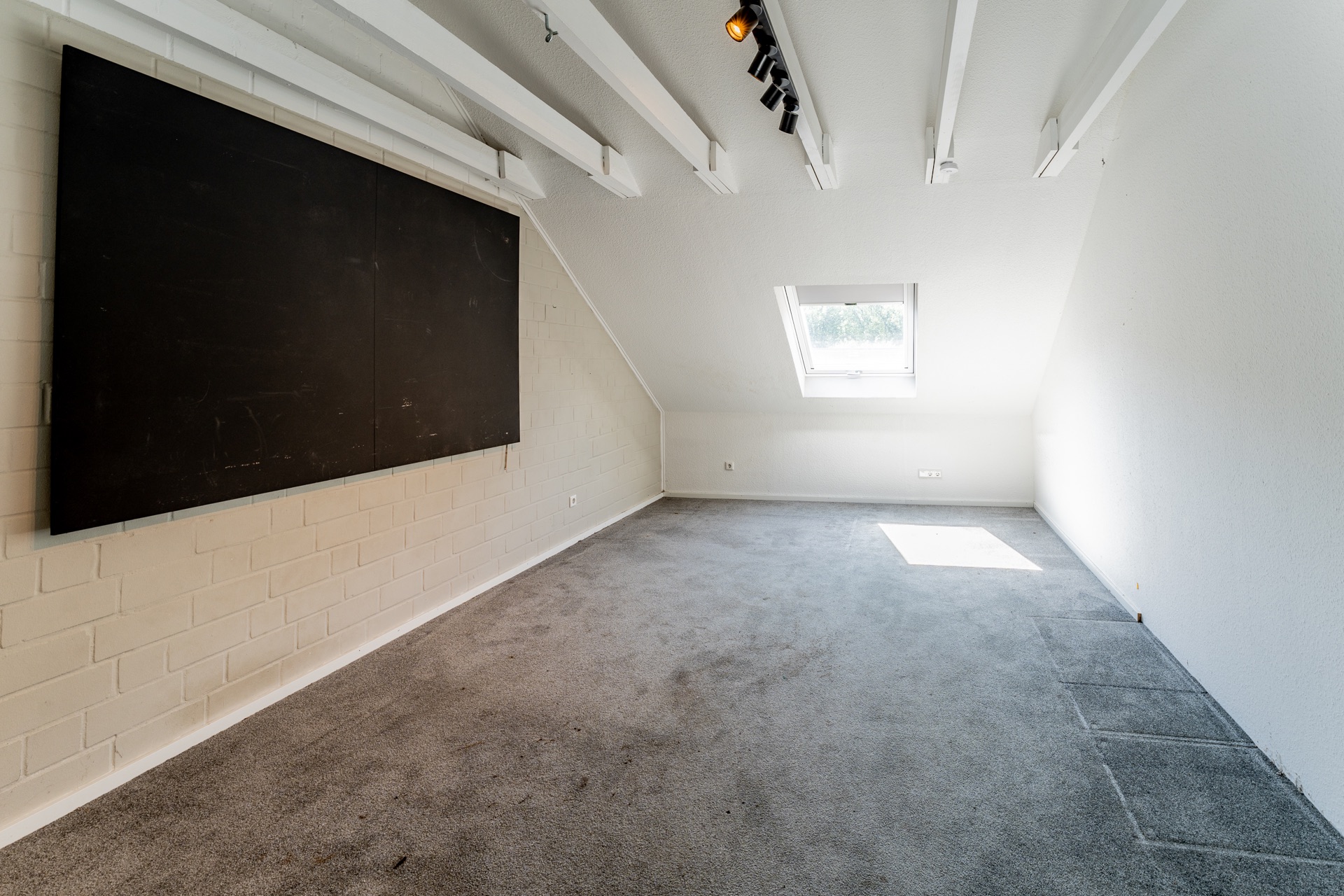 Heller Dachbodenraum mit Teppich, Schrägfenster und großer Tafel an der Wand.