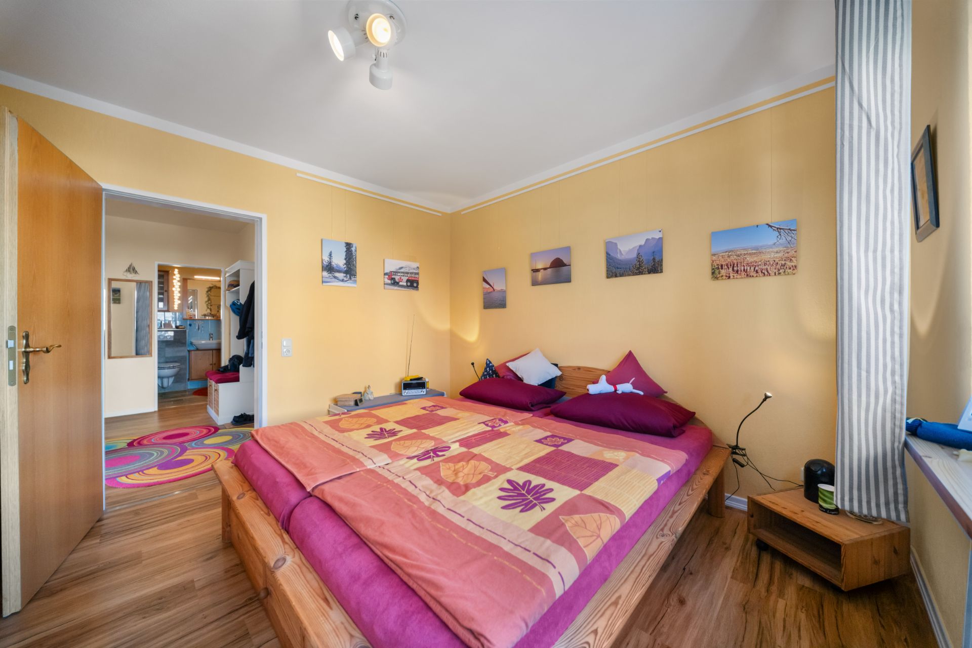 Schlafzimmer mit Doppelbett, gelben Wänden, Bildern an der Wand und offen stehender Tür zum Flur.