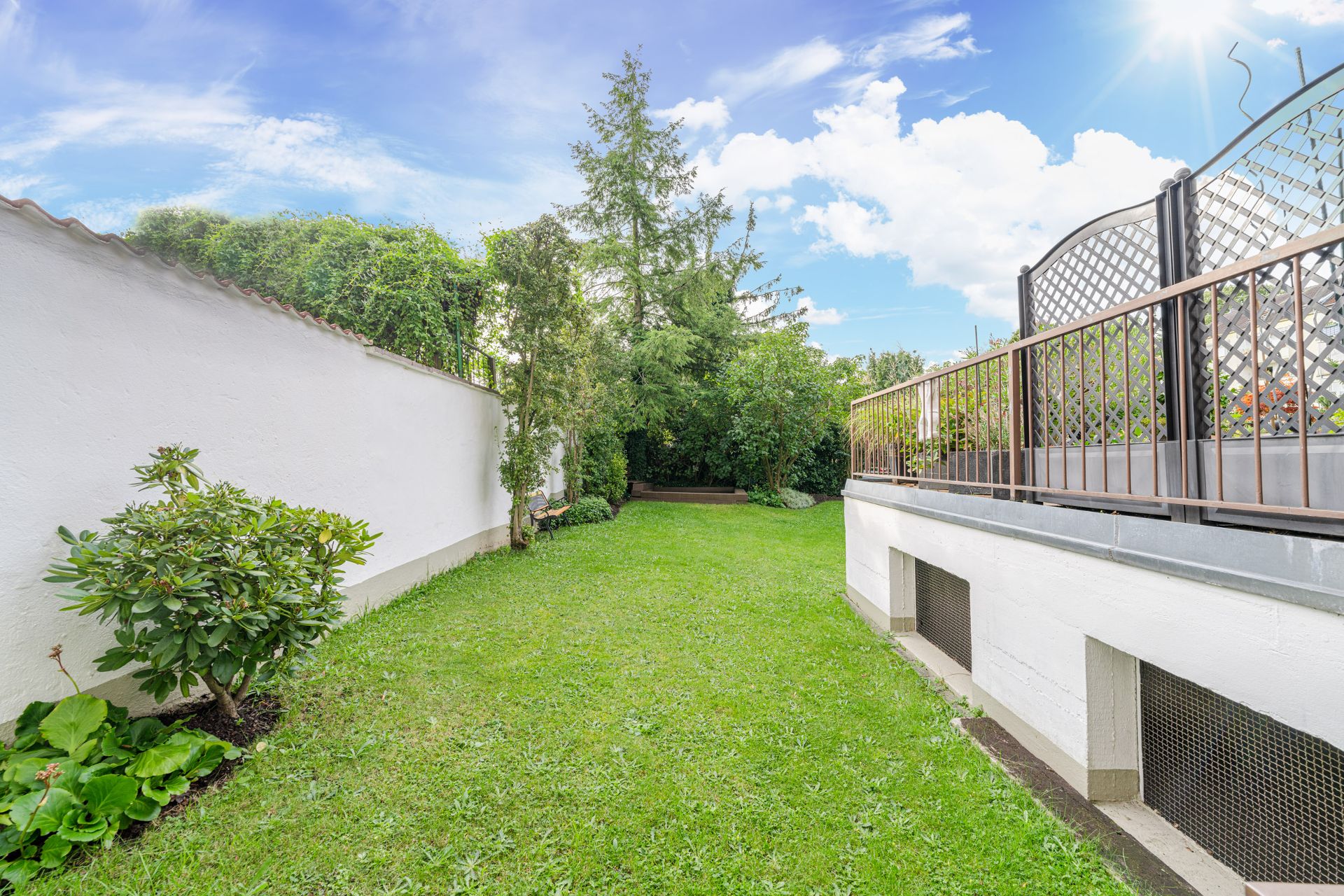 Grüner Garten mit Rasen, Sträuchern, Bäumen, weißer Mauer und Terrasse mit Gitterzaun.