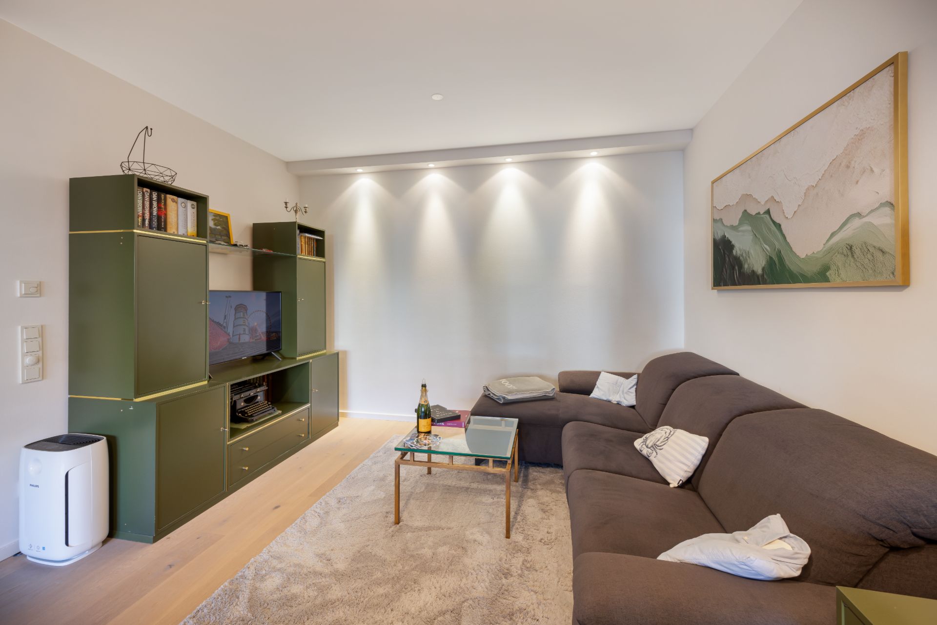 Wohnzimmer mit braunem Sofa, grünem Regal, Teppich und Wandbild.
