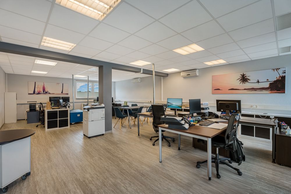 Großer heller Raum mit mehreren Schreibtischen, Bürostühlen und Computern, Bilder an den Wänden.
