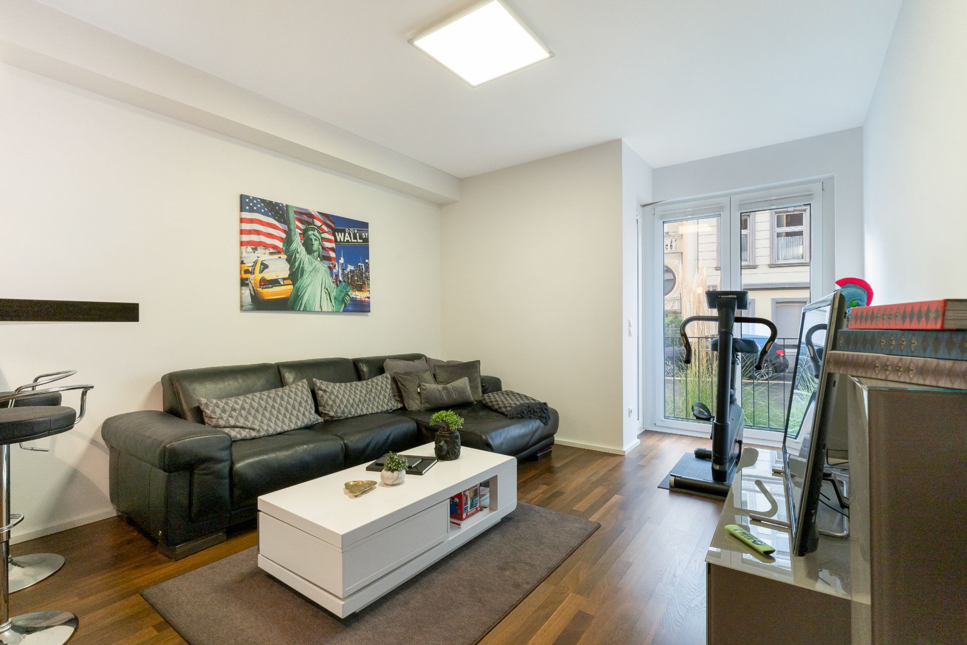 Modernes Wohnzimmer mit schwarzem Sofa, Couchtisch, Fernseher und Fitnessgerät vor Fenster.