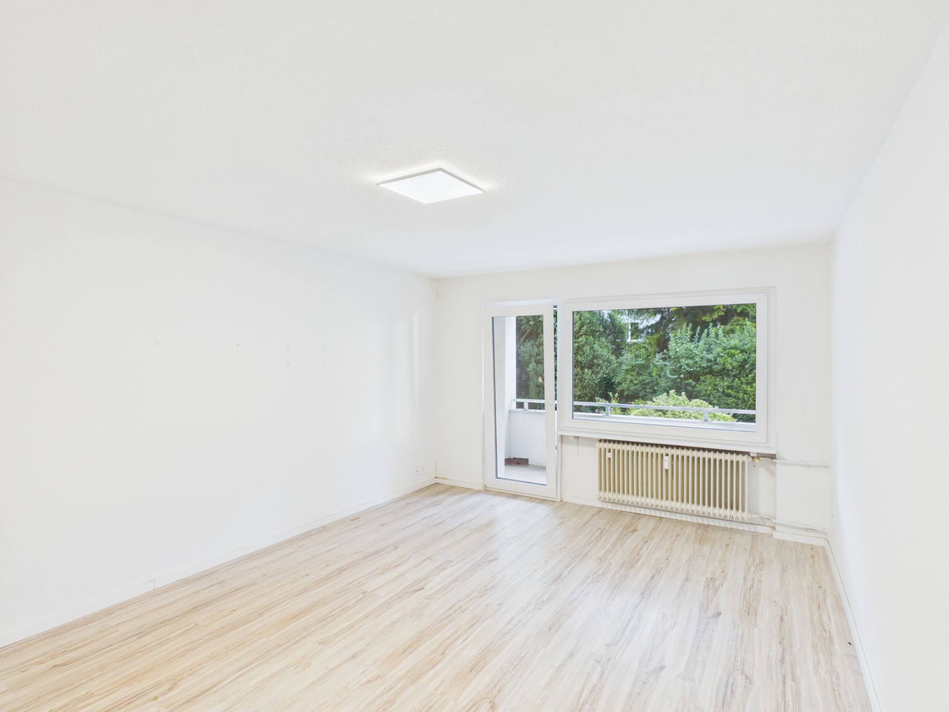 Helles, leeres Zimmer mit Holzfußboden, großem Fenster und Tür zu einem Balkon.