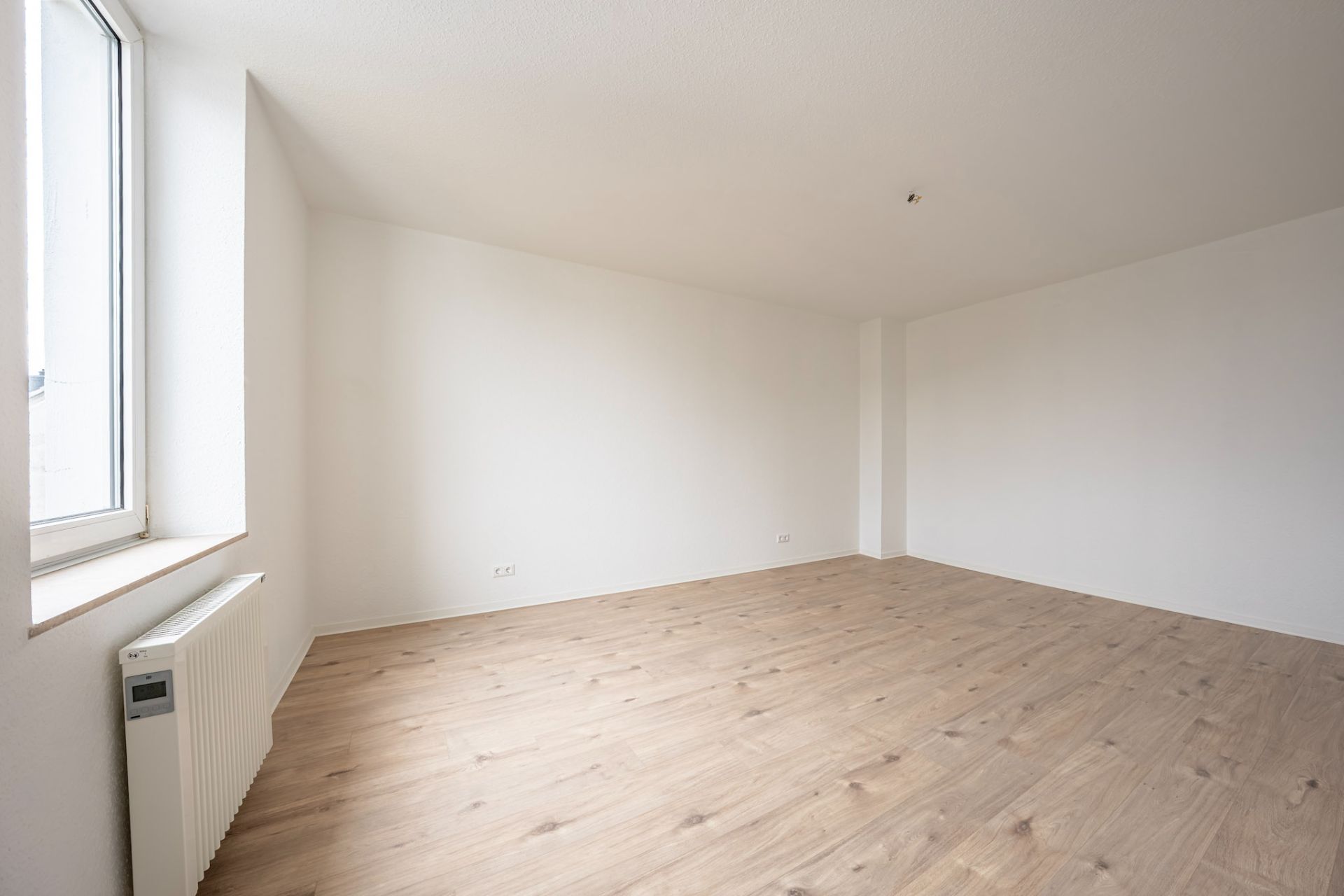 Leerer Raum mit hellem Holzboden, weißer Wand und großem Fenster links.