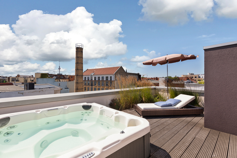 Dachterrasse mit Whirlpool, Sonnenliege, Sonnenschirm und Stadtblick bei klarem Himmel.