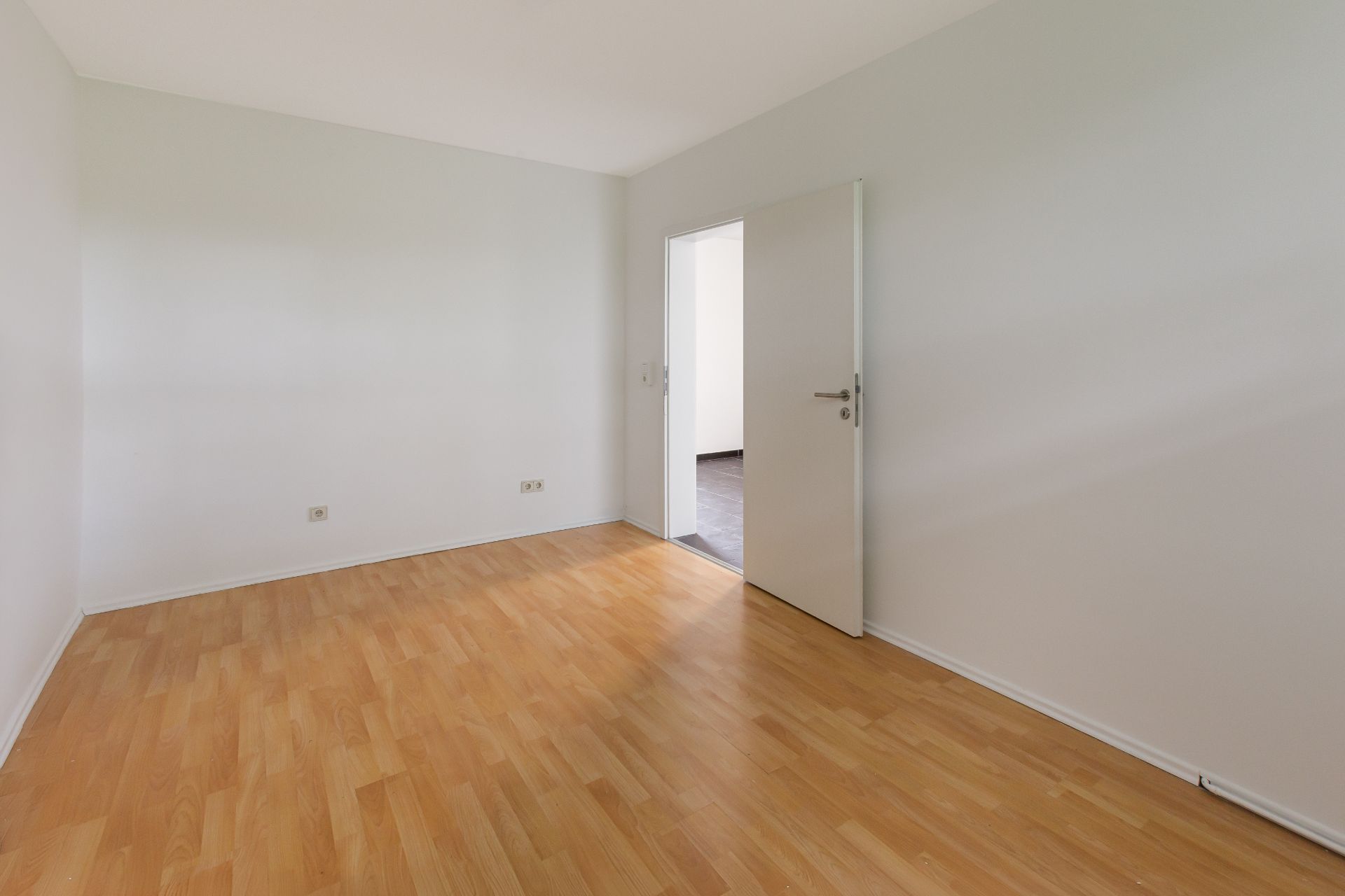 Leerer Raum mit hellem Holzboden, weißen Wänden und offener Tür.
