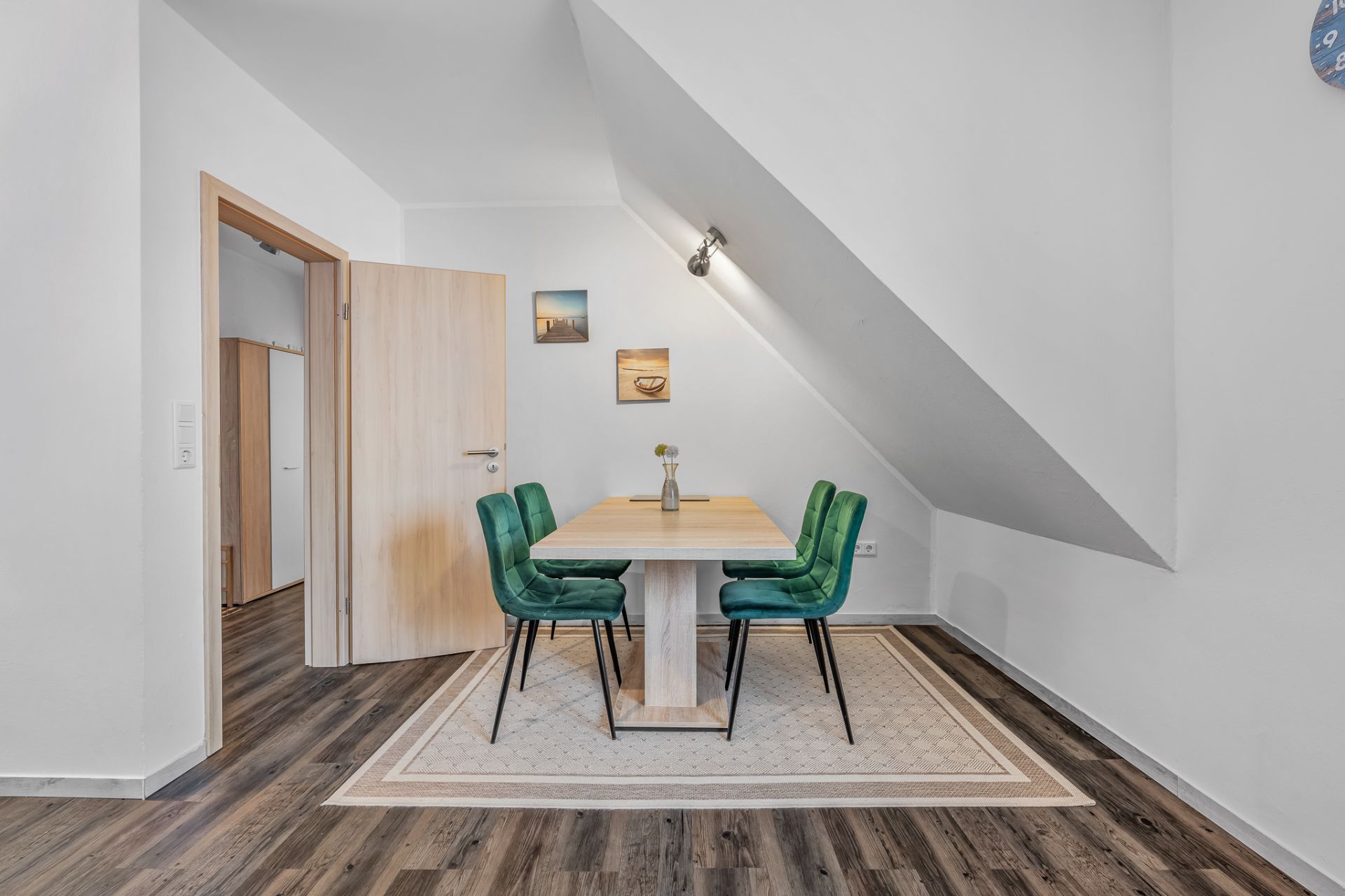 Essbereich mit Holztisch, vier grünen Stühlen und schrägem Dach, Holzboden und Wandbildern.