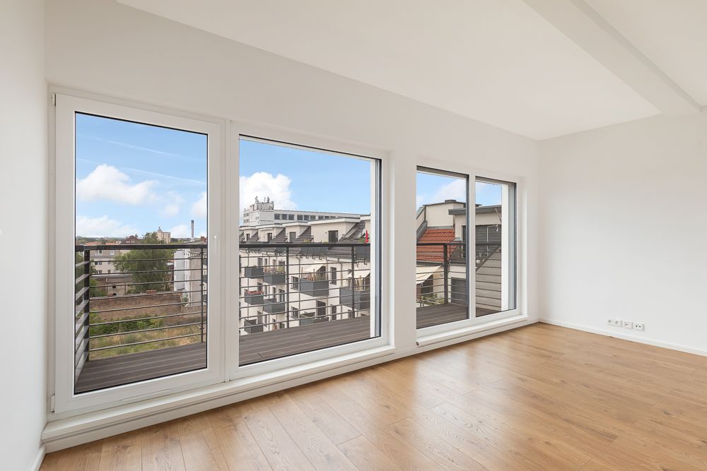 Helles, leeres Zimmer mit Holzfußboden und großen Fenstern mit Balkonblick auf Stadtgebäude.