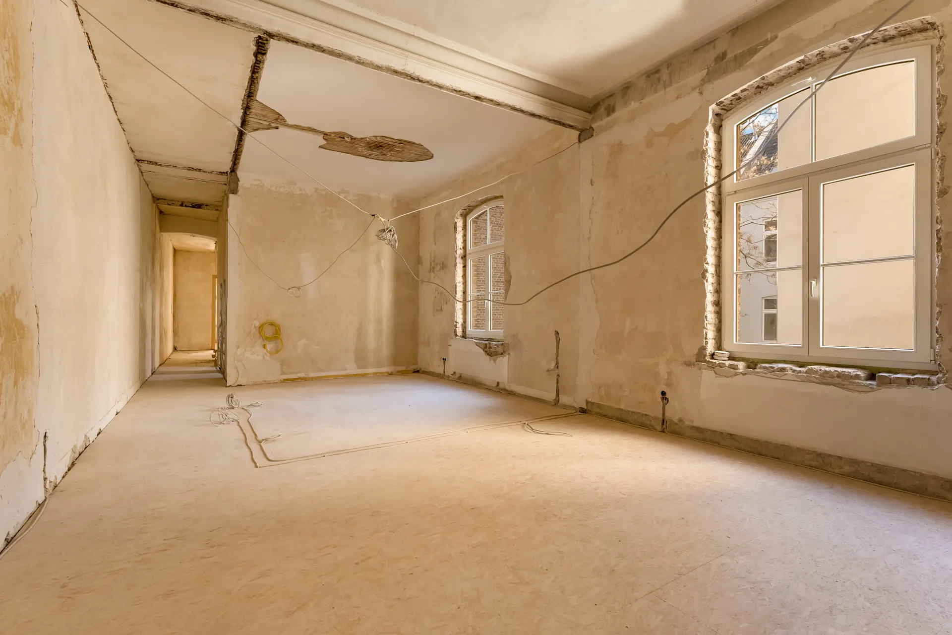 Leerer Raum mit renovierungsbedürftigen Wänden, offenliegenden Kabeln und großen Fenstern.