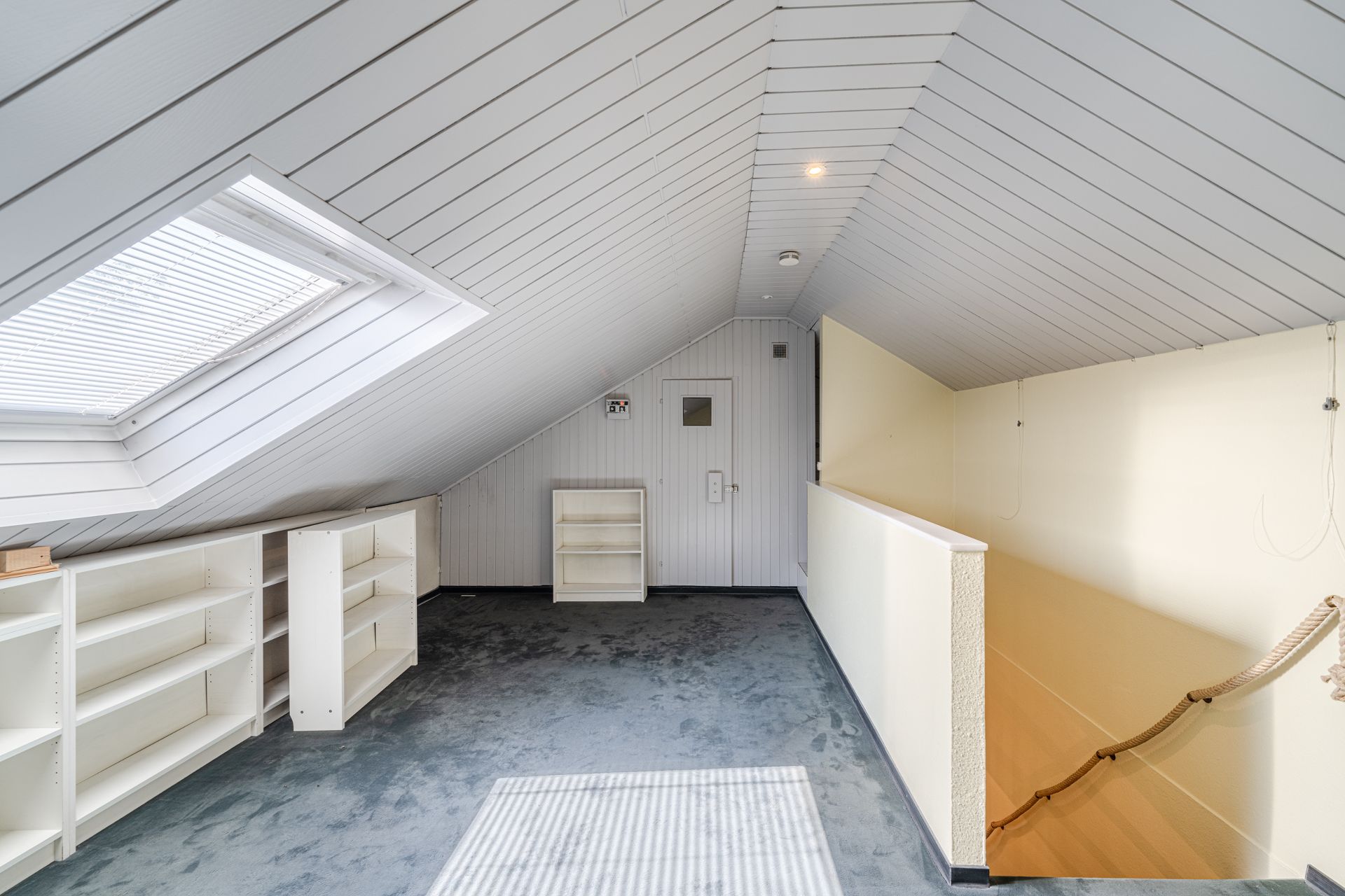 Dachgeschossraum mit Schräge, großem Fenster, Regalen und Treppe mit Seilgeländer.