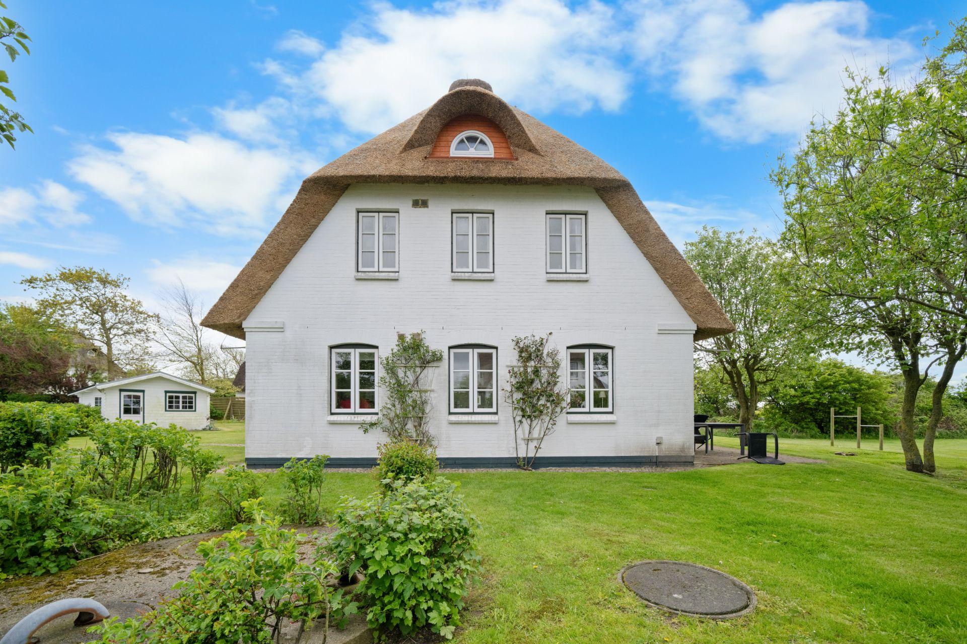 Weißes Haus mit Reetdach im Garten, umgeben von grüner Wiese und Bäumen.
