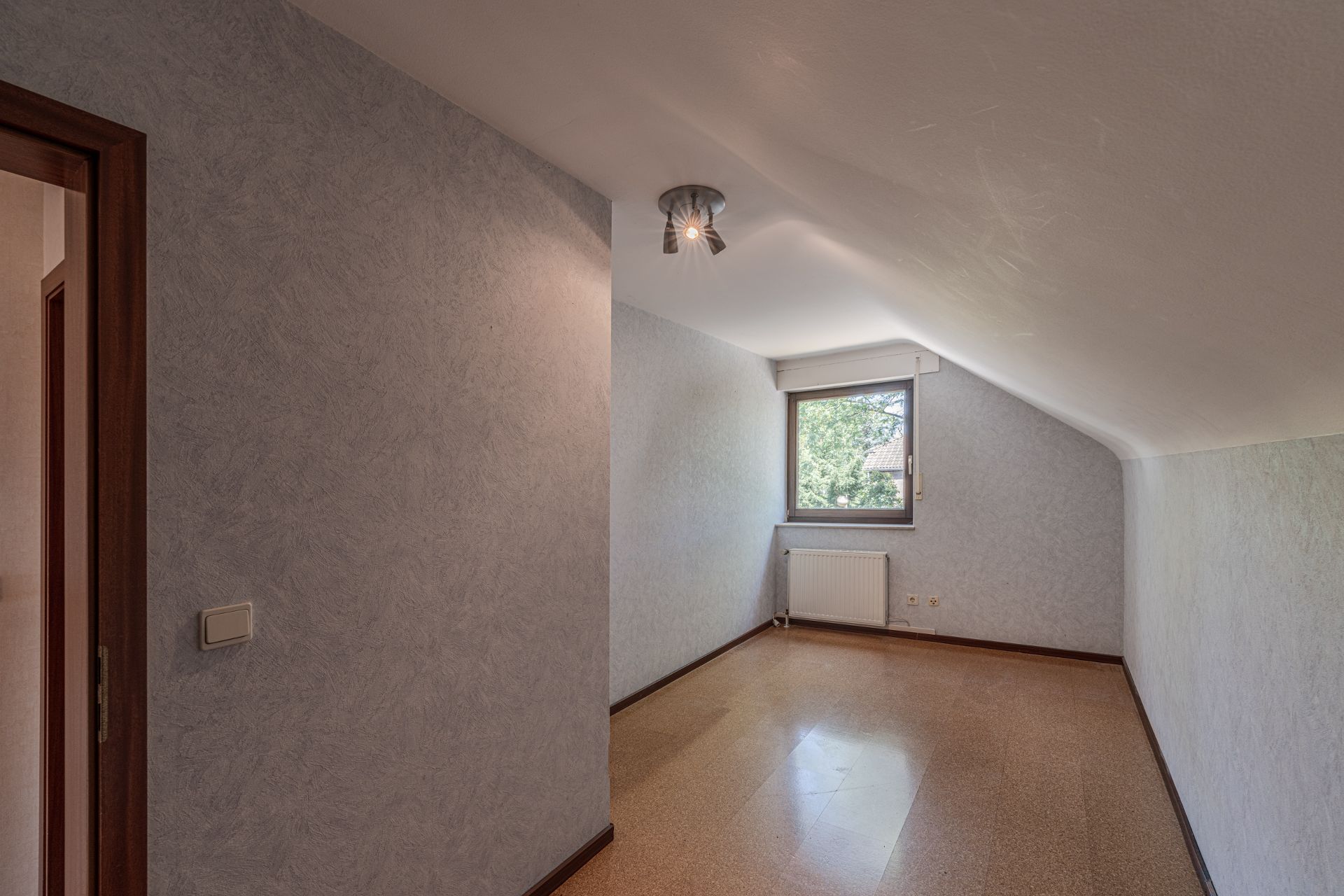 Schmaler Raum mit schräger Decke, Fenster, Heizkörper und hellen Wänden.