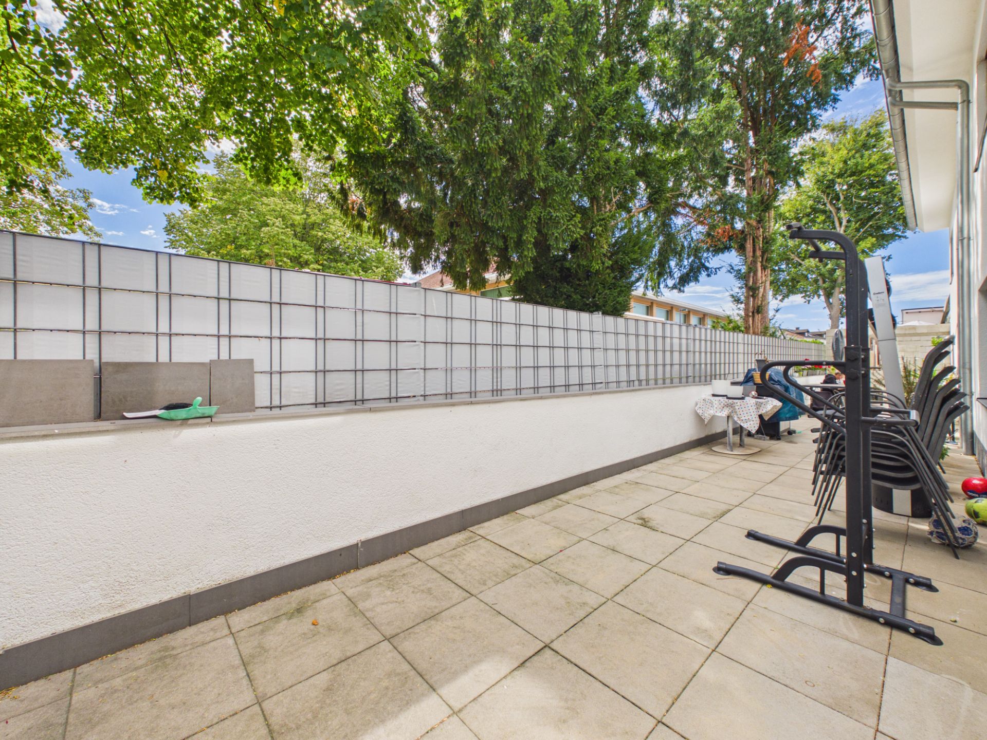 Geflieste Terrasse mit Fitnessgerät, Stühlen und Tisch, daneben Bäume und hoher Sichtschutzzaun.