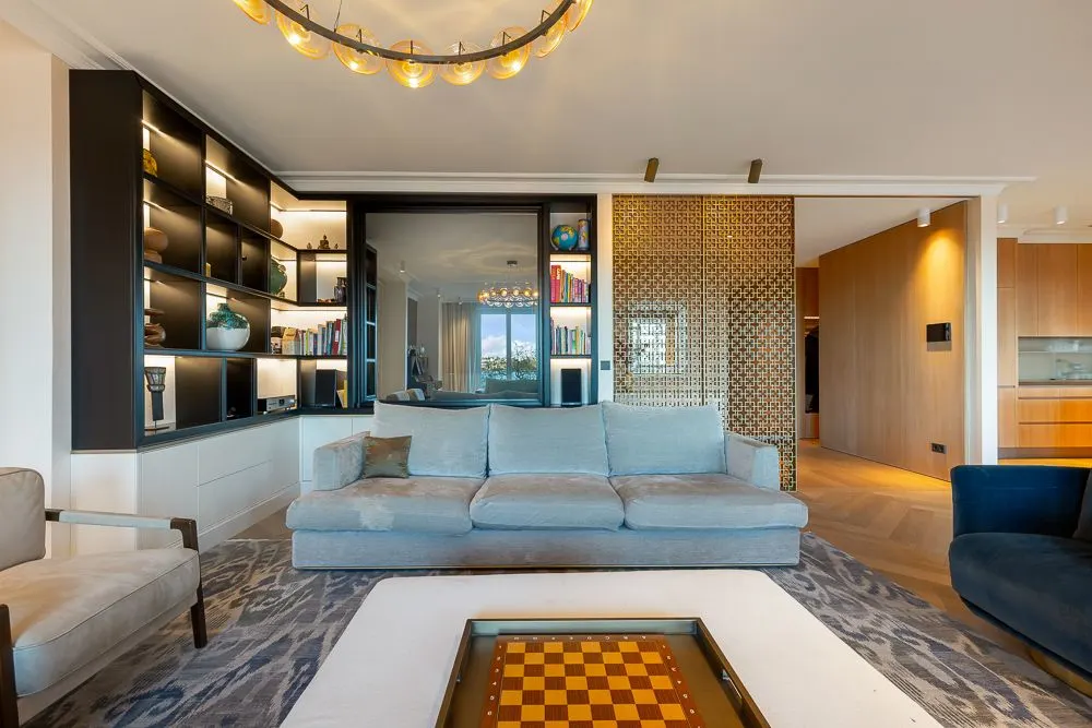 Modernes Wohnzimmer mit grauem Sofa, Bücherregal, Schachbrett und heller Beleuchtung.