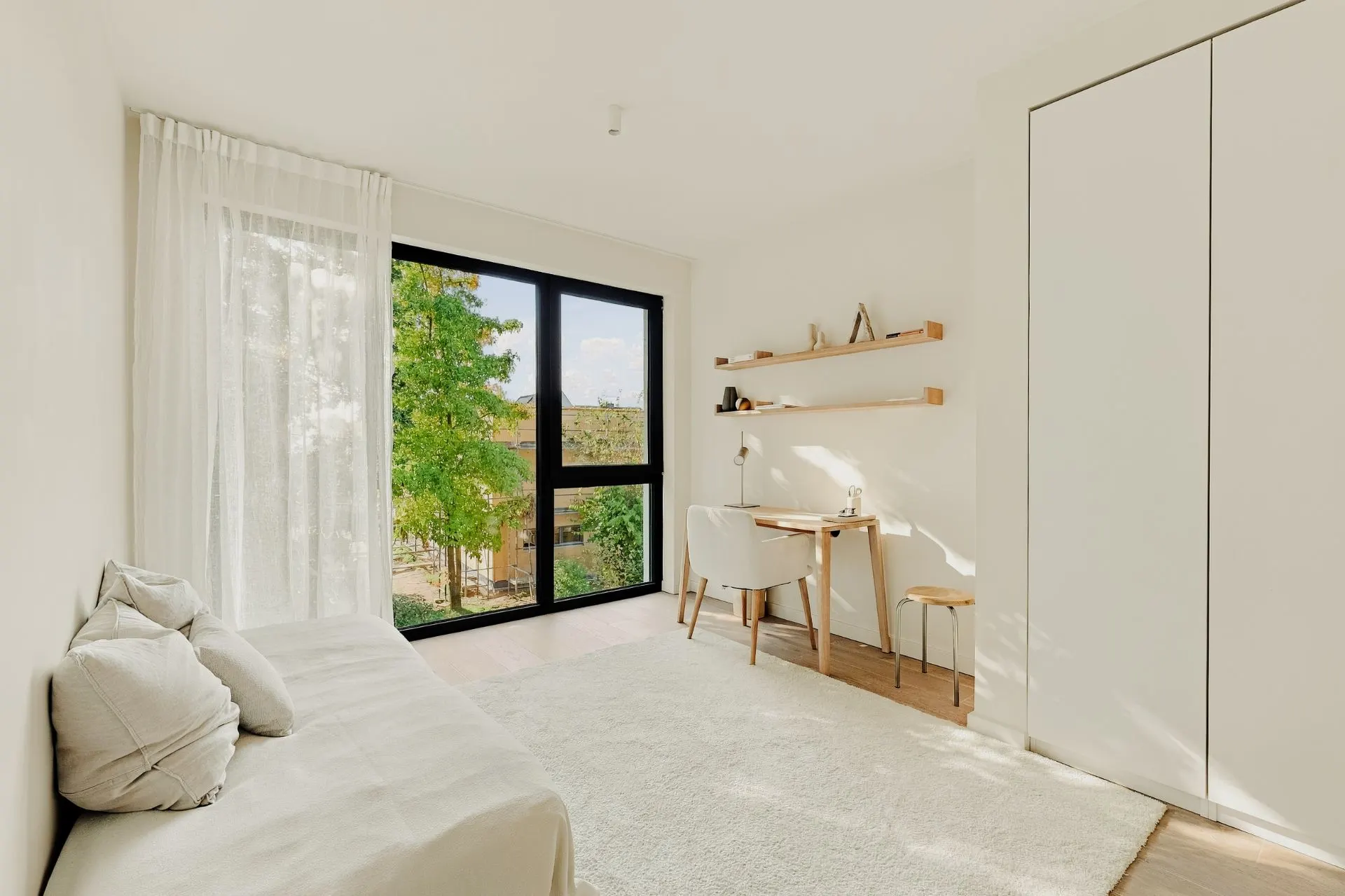 Helles Zimmer mit Sofa, Schreibtisch, Regalen, großem Fenster und weißem Schrank.