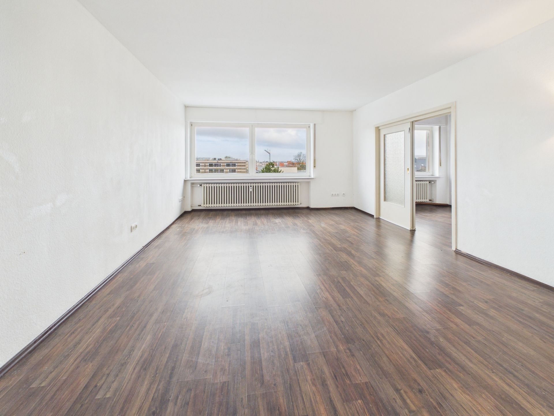 Leeres Wohnzimmer mit großem Fenster, weißer Wand und dunklem Holzboden.