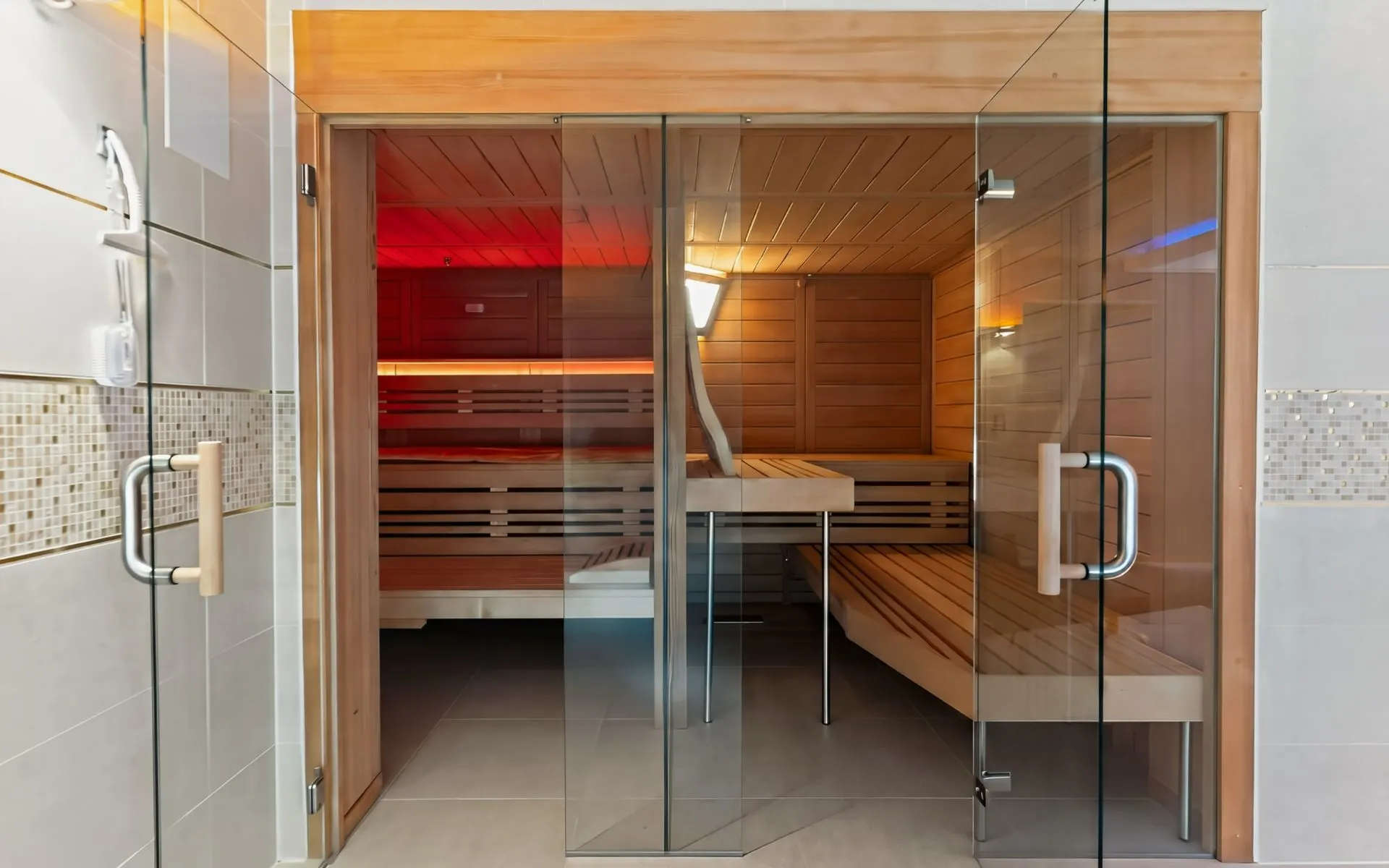 Moderne Sauna mit Holzbänken und Glastür, beleuchtet von Wandlampen.