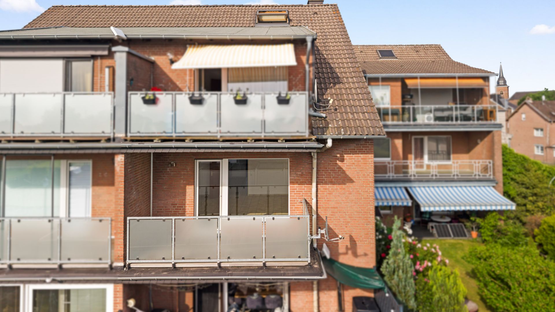 Mehrfamilienhaus mit Balkonen, Glasgeländern und Markisen, umgeben von Garten.