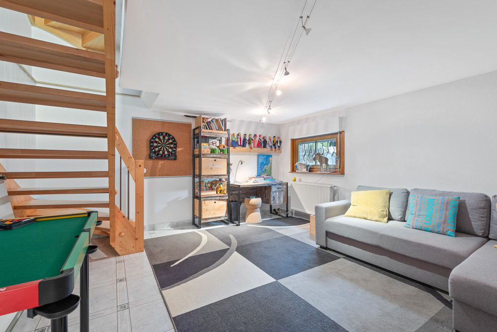 Heller Kellerraum mit grauem Sofa, Schreibtisch, Regal, Teppich und Holztreppe.