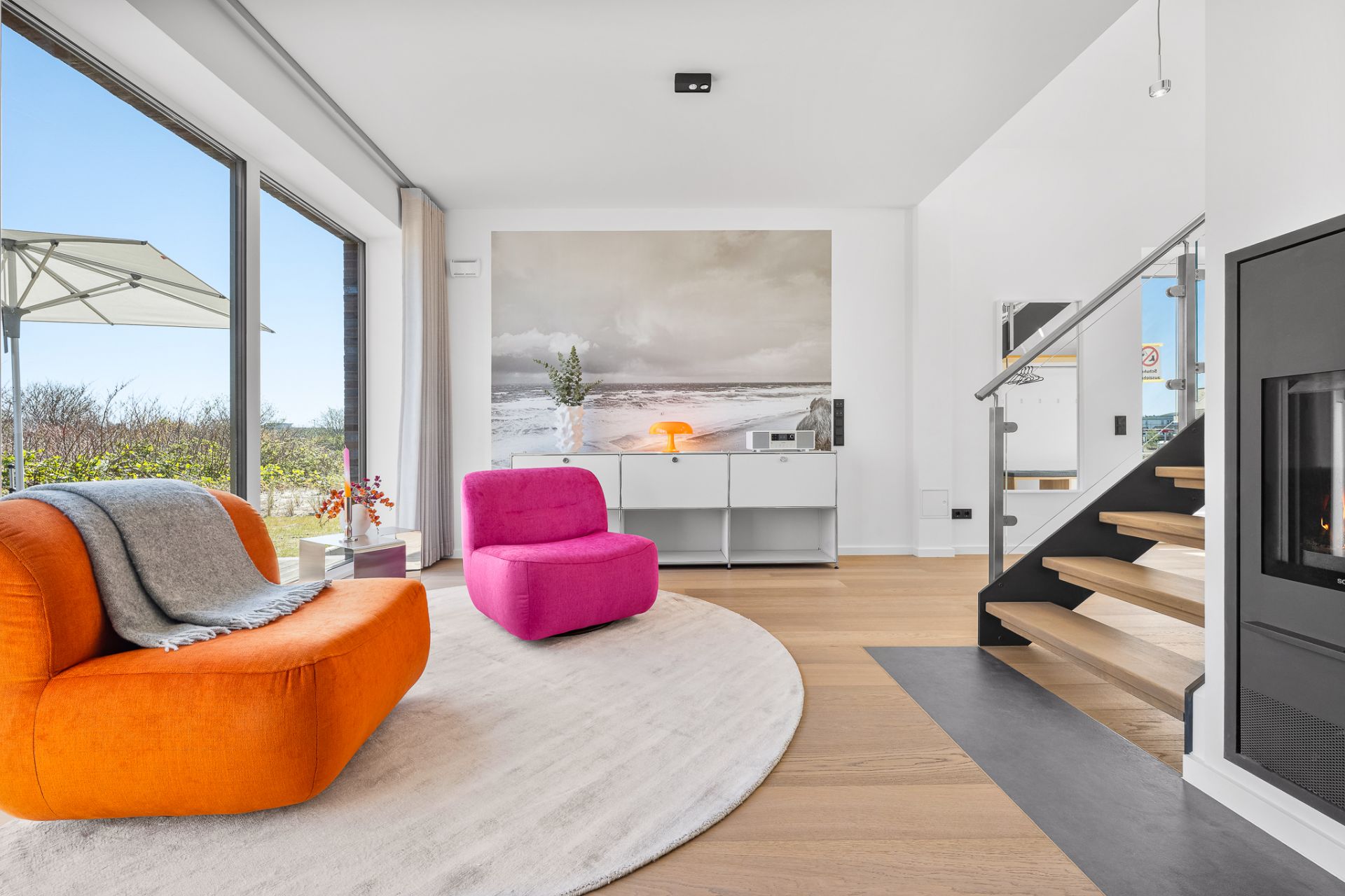 Modernes Wohnzimmer mit großen Fenstern, orange und pinkem Sessel, Kamin und Treppe.