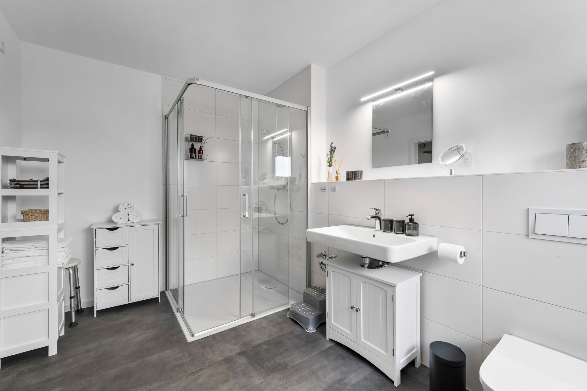 Modernes Bad mit Dusche, Waschbecken, Spiegel und weißen Möbeln auf grauem Boden.