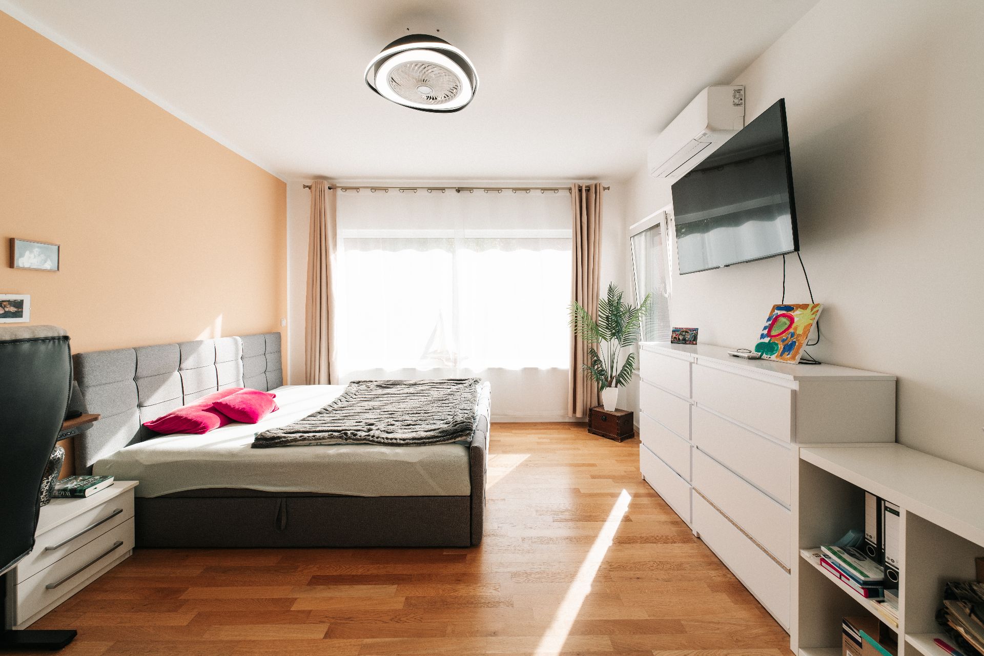 Helles Schlafzimmer mit Doppelbett, Kommoden, großem Fenster und Wandfernseher.