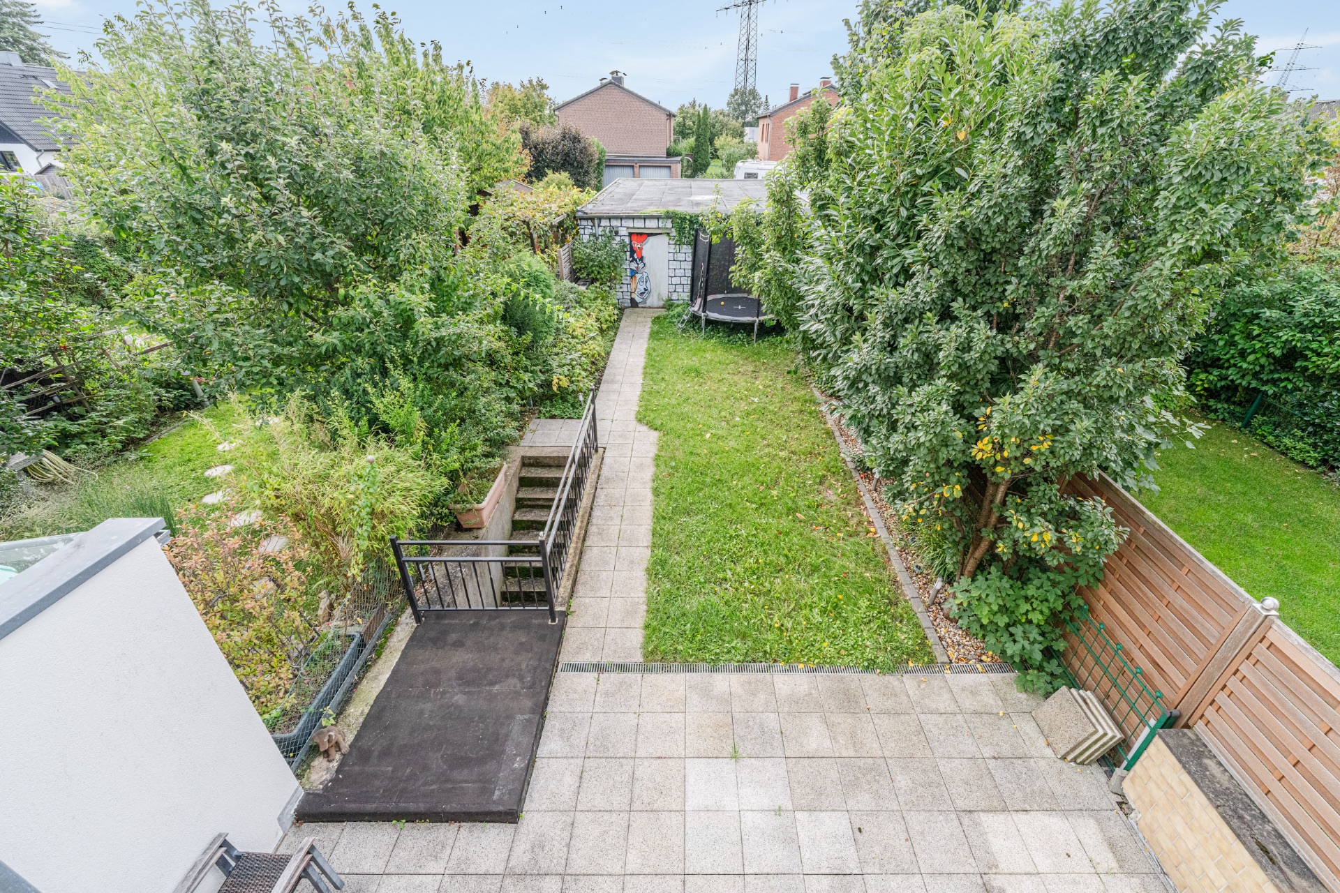 Blick auf einen Garten mit Rasenfläche, Terrasse, Weg, Bäumen und Sträuchern.