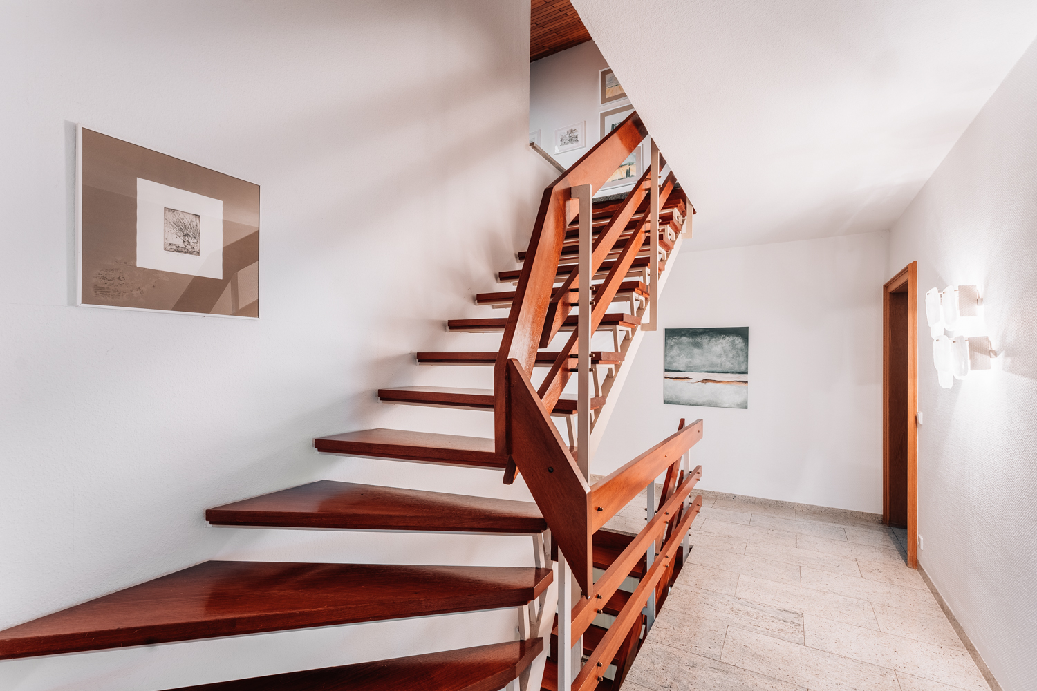 Innenbereich mit moderner Holztreppe, weißer Wand und Bildern im Flur.