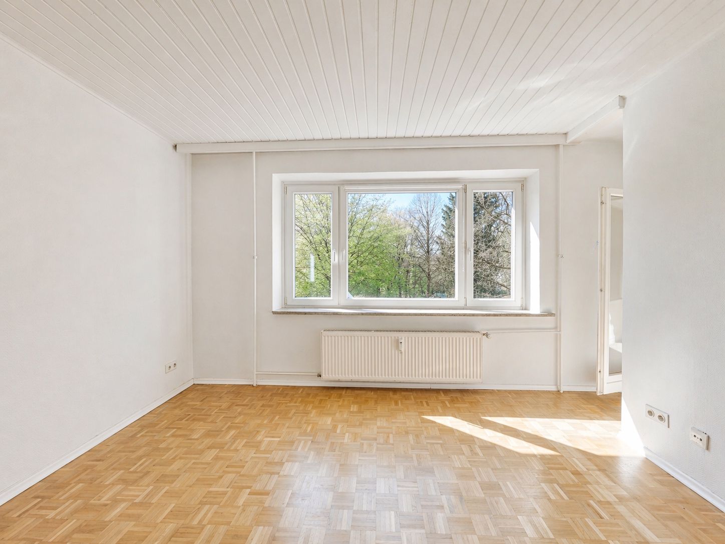 Helles, leeres Zimmer mit Parkettboden, großem Fenster und Heizkörper darunter.