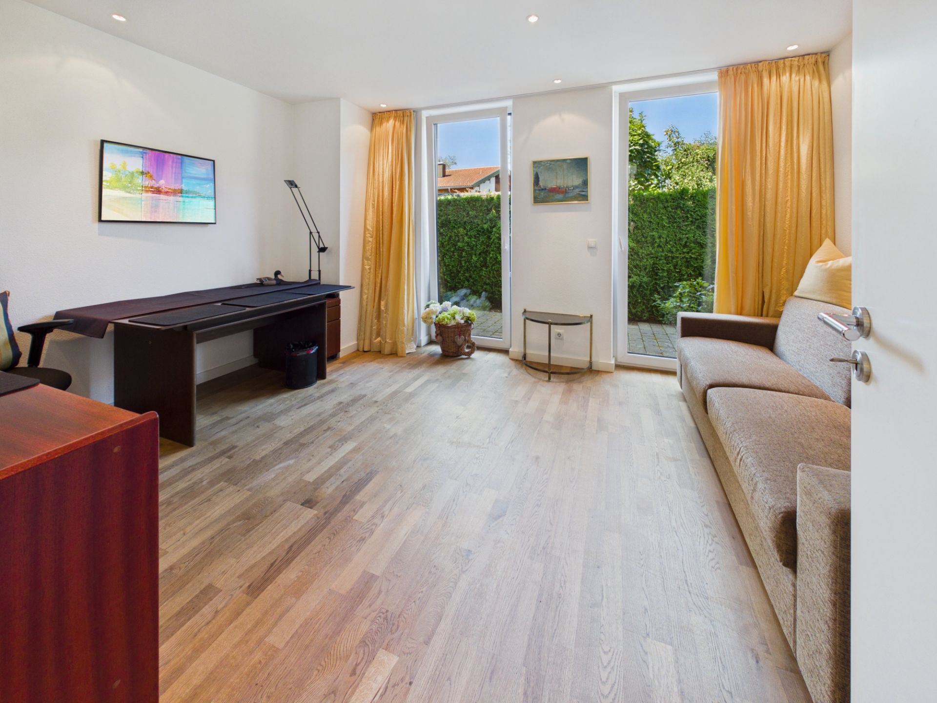 Helles Zimmer mit Holzboden, Sofa, Schreibtisch, großen Fenstern und gelben Vorhängen.