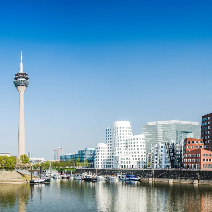 Stadtsilhouette mit Fluss, modernen Gebäuden und einem hohen Fernsehturm unter blauem Himmel.