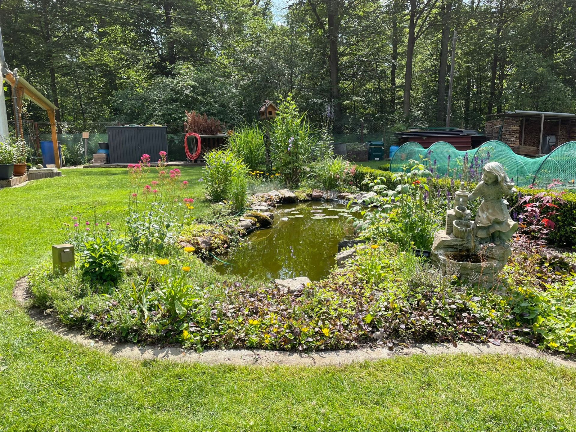 Garten mit kleinem Teich, Blumen und einer Steinfigur eines Mädchens.