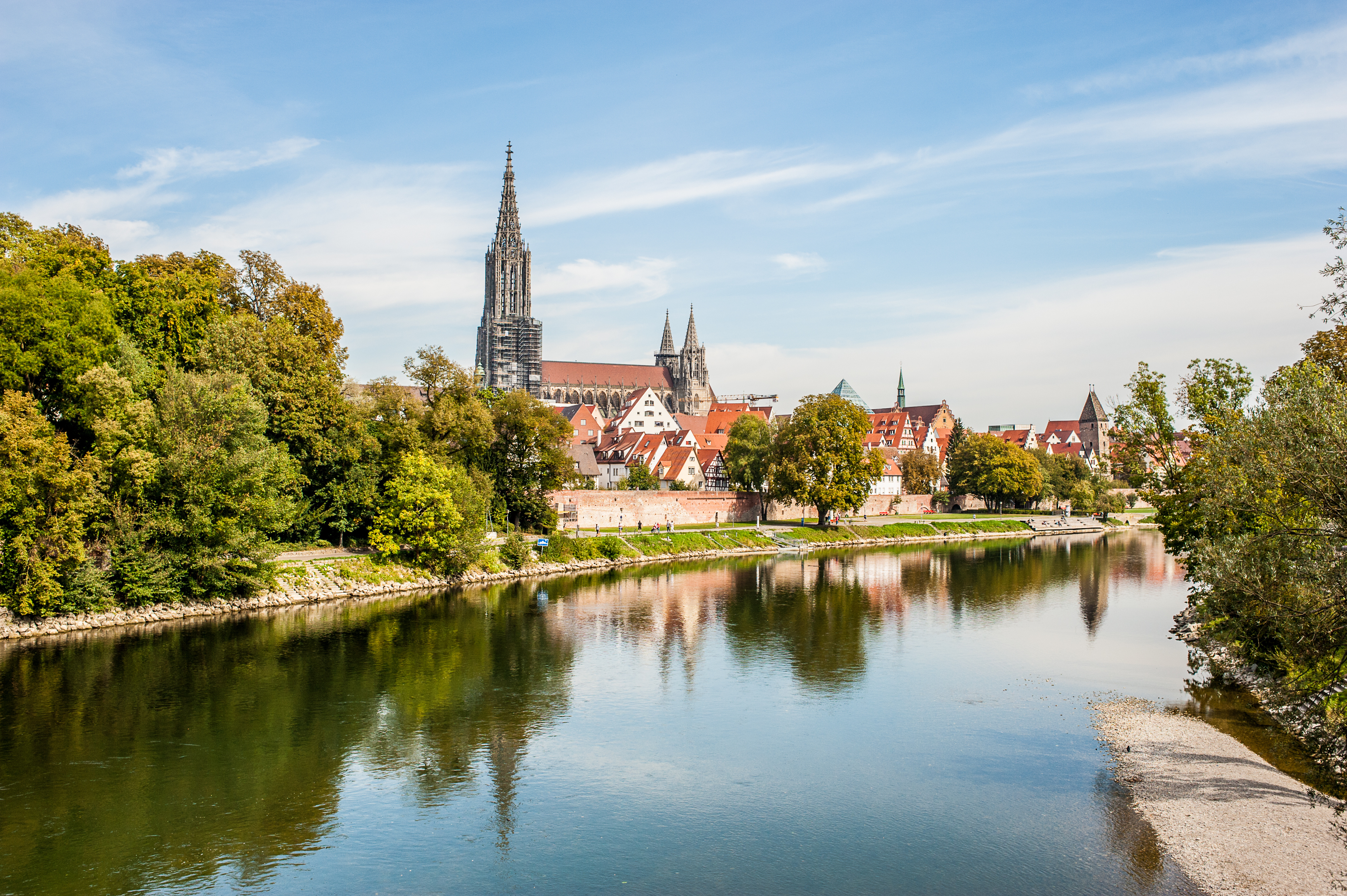 Stadtansicht mit Fluss im Vordergrund, Bäumen und Kirchturm im Hintergrund.