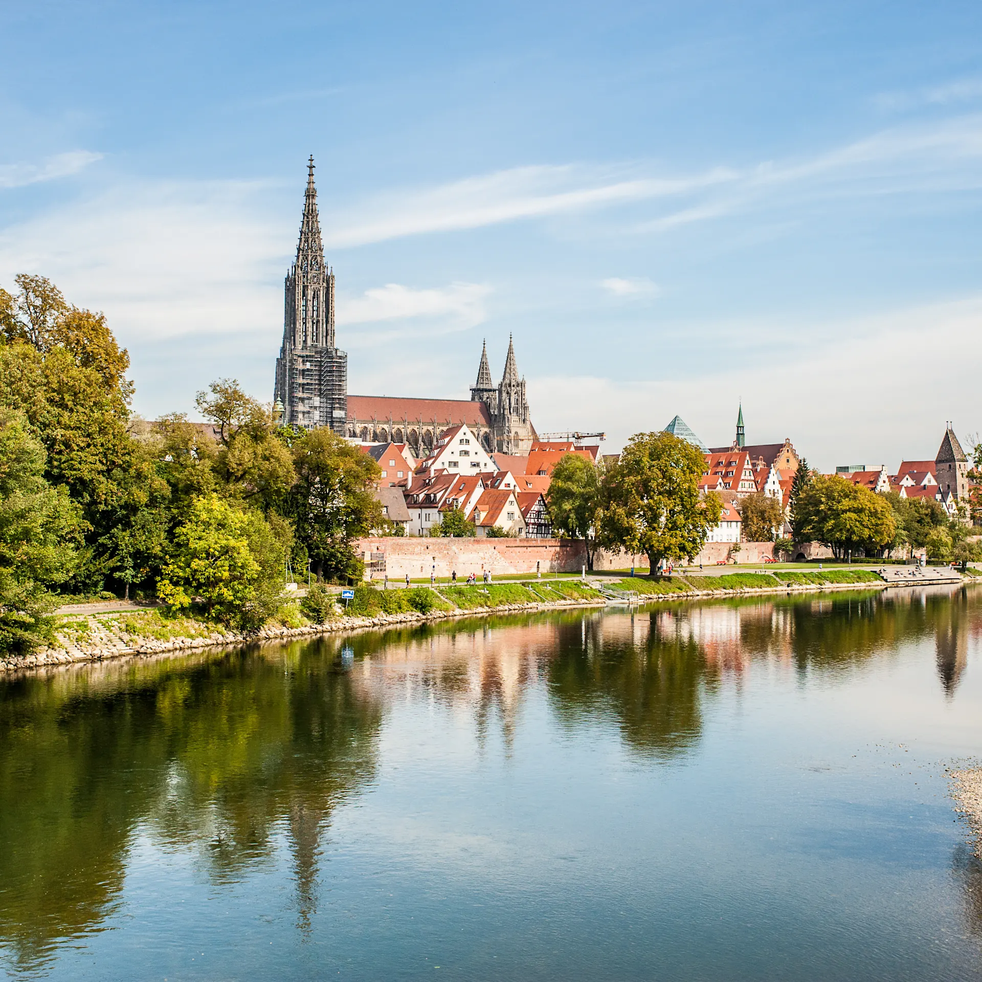 Stadtansicht mit Fluss im Vordergrund, Bäumen und Kirchturm im Hintergrund.