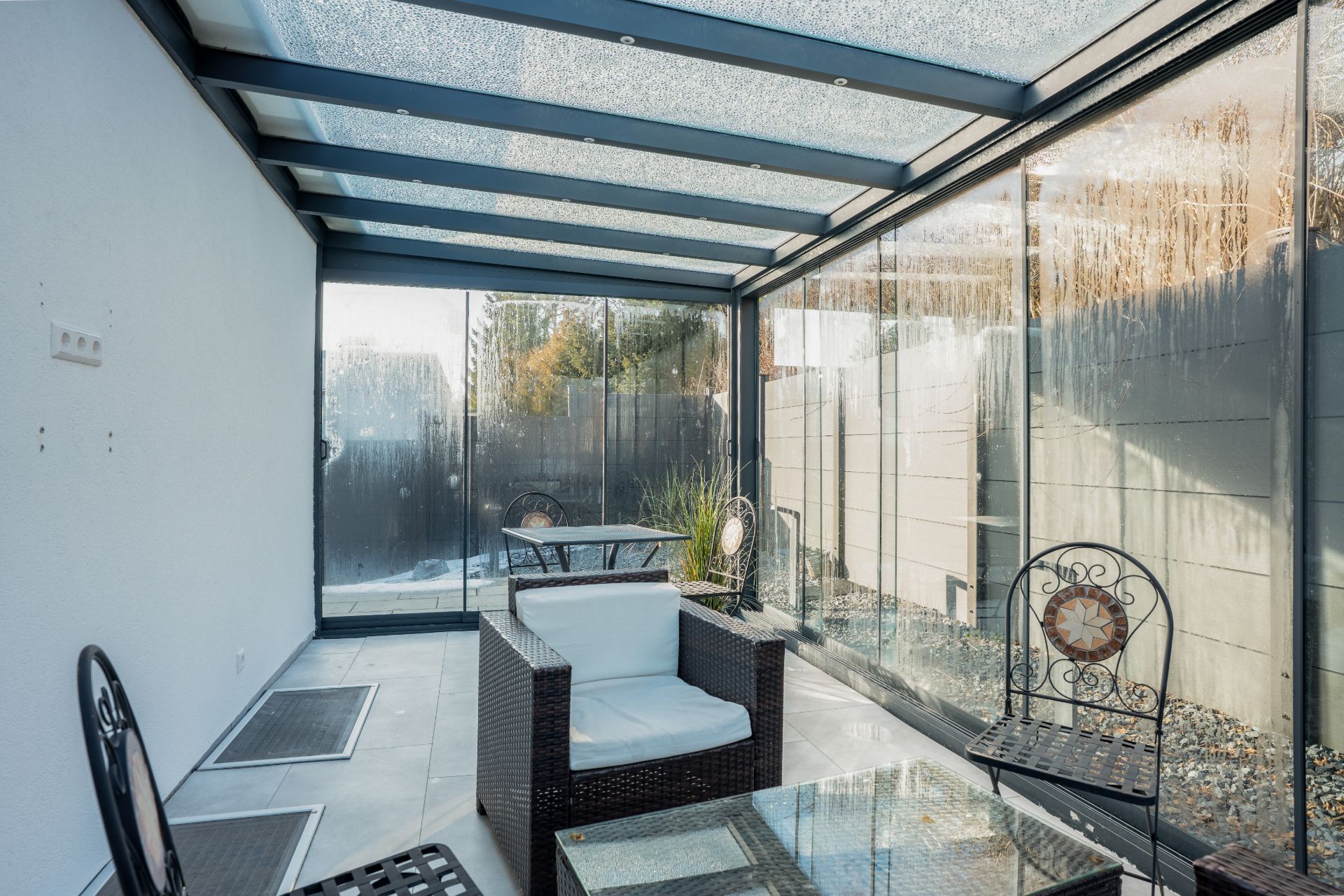 Überdachte Terrasse mit Glaswänden, Sitzmöbeln und Tisch auf Fliesenboden.