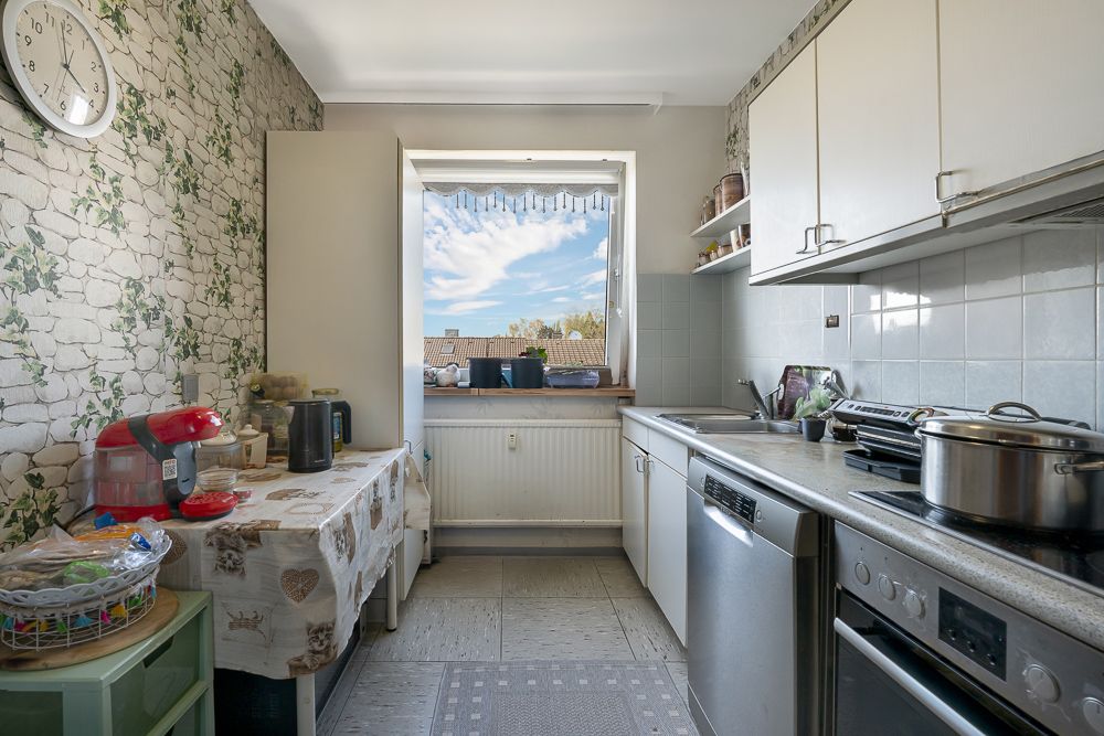 Schmale Küche mit Fenster, weißen Schränken, Tischdecke und Geräten auf der Arbeitsfläche.
