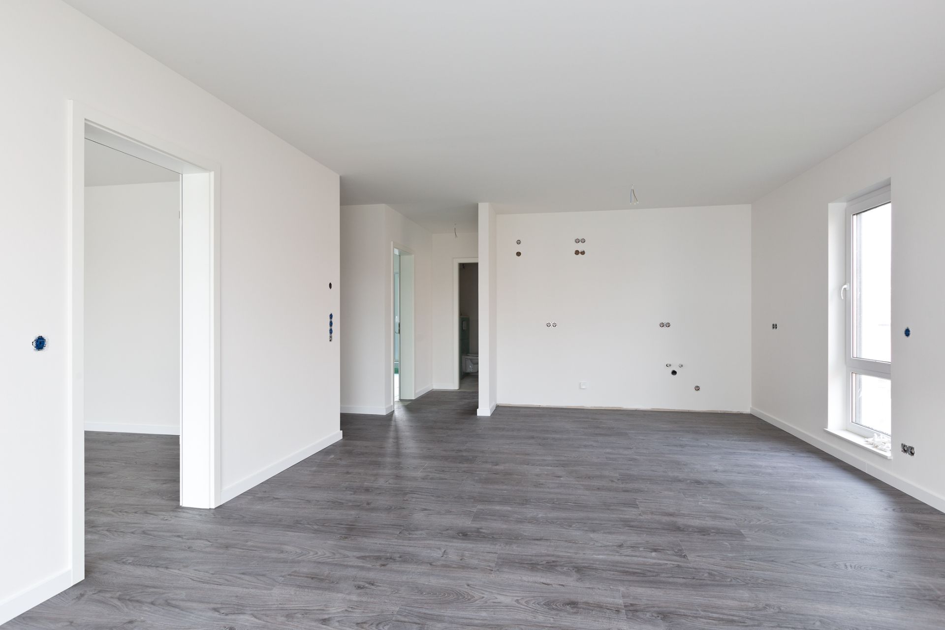 Leerer, heller Raum mit grauem Boden und weißen Wänden, mehrere Türen und Fenster sichtbar.