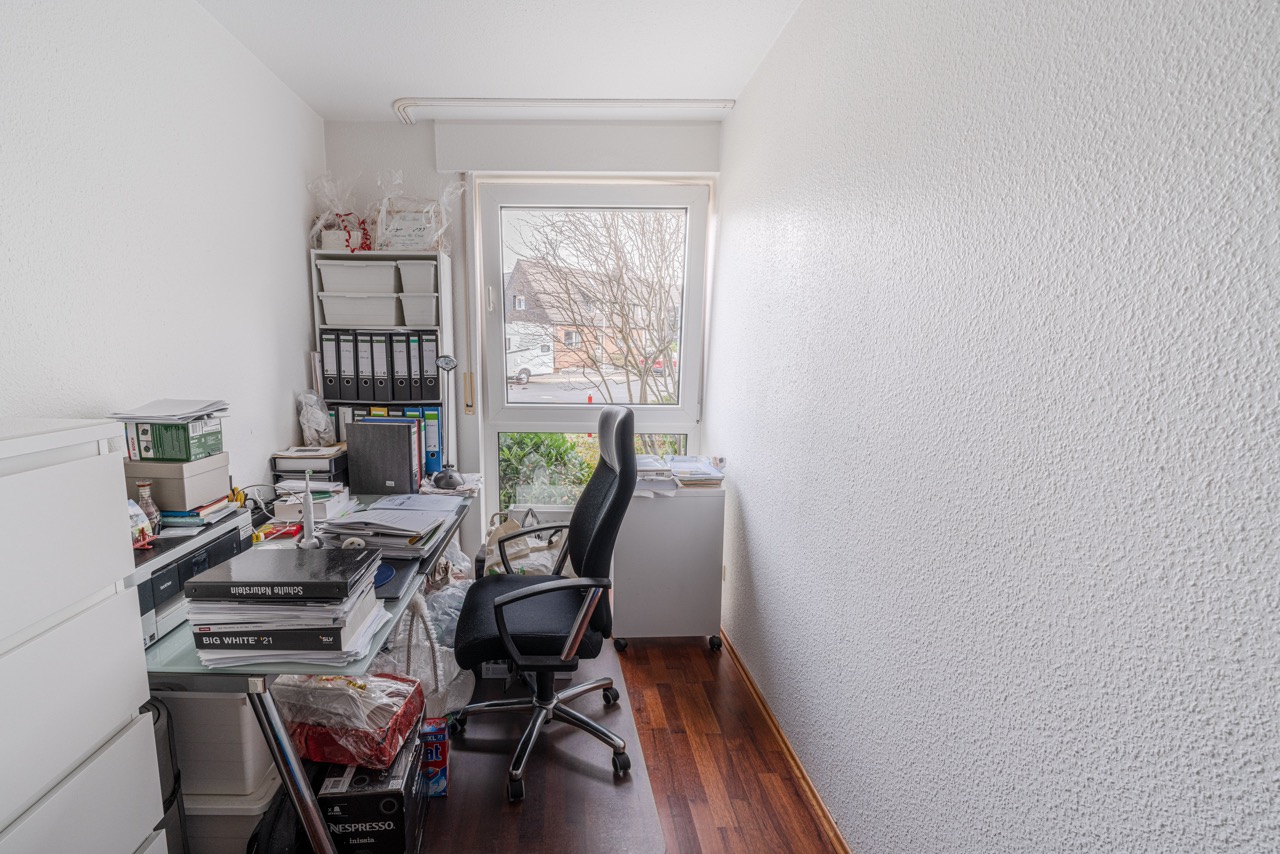 Kleines, vollgestelltes Arbeitszimmer mit Schreibtisch, Bürostuhl und Regal vor einem Fenster.