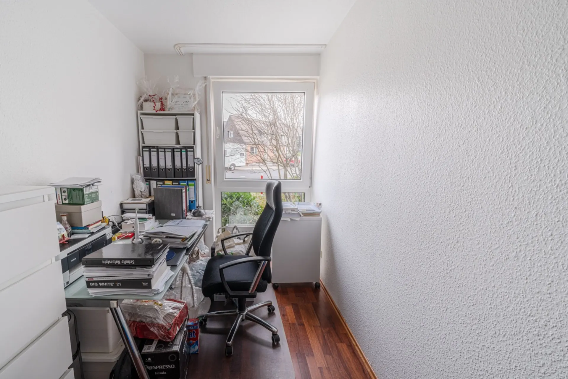 Kleines, vollgestelltes Arbeitszimmer mit Schreibtisch, Bürostuhl und Regal vor einem Fenster.