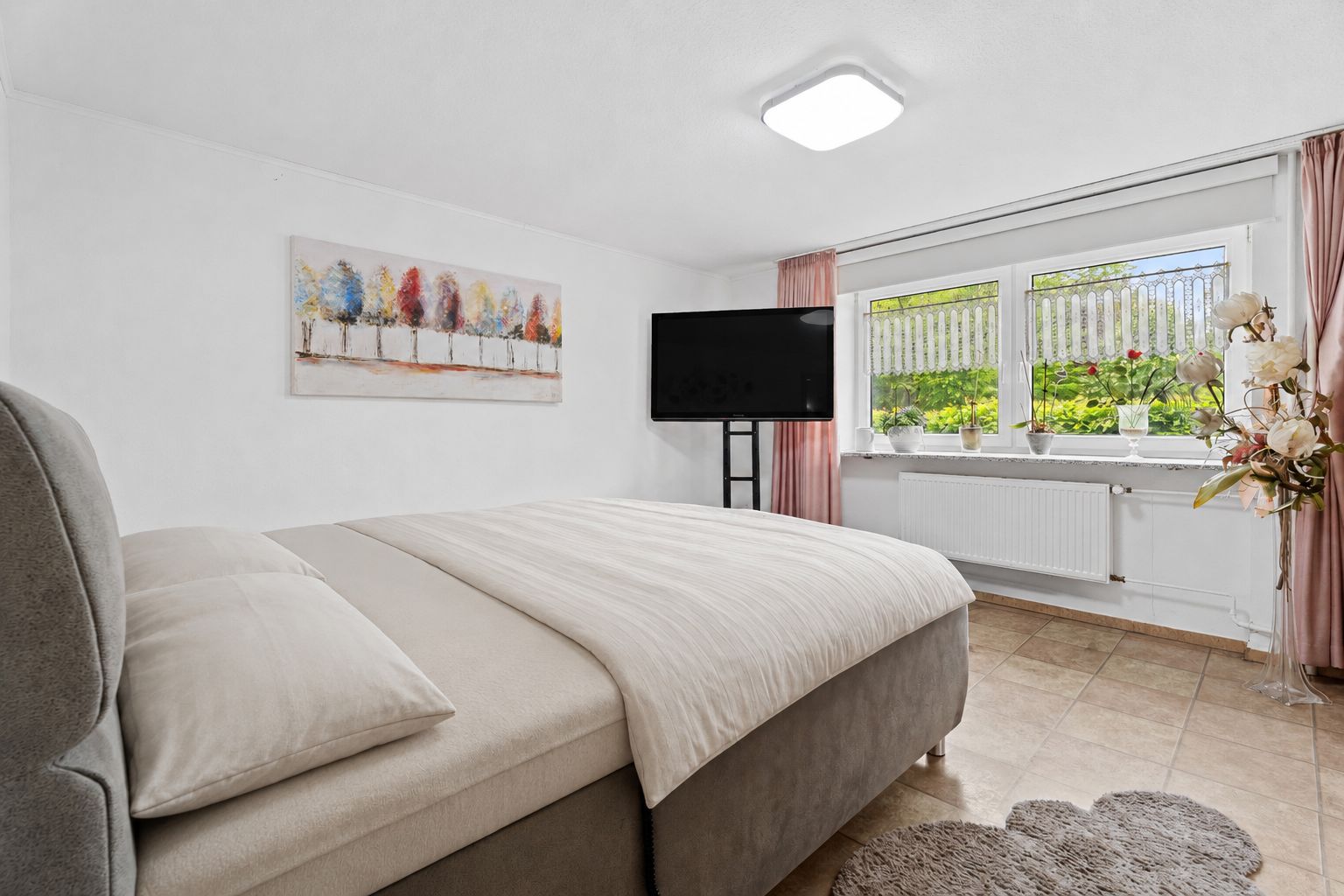 Helles Schlafzimmer mit Doppelbett, großem Fenster, TV und Dekorationen.