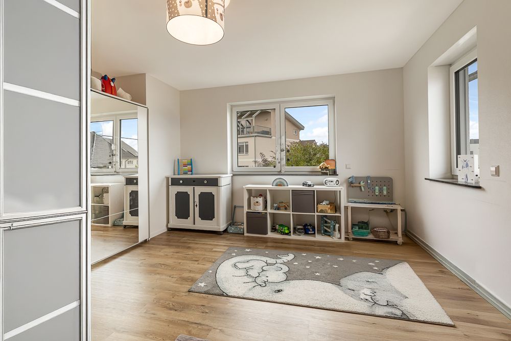 Helles Kinderzimmer mit Spielteppich, Regal und Fenster, Holzboden und weißer Wand.