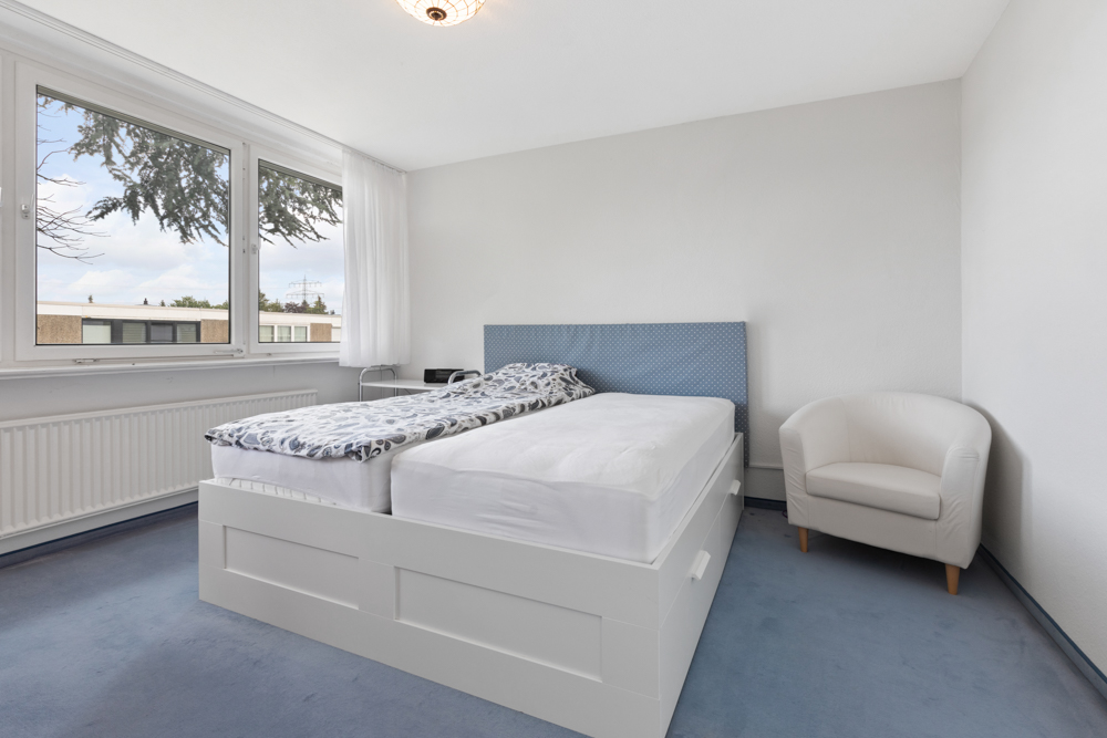 Helles Schlafzimmer mit großem Bett, blauem Teppich und weißem Sessel neben dem Fenster.