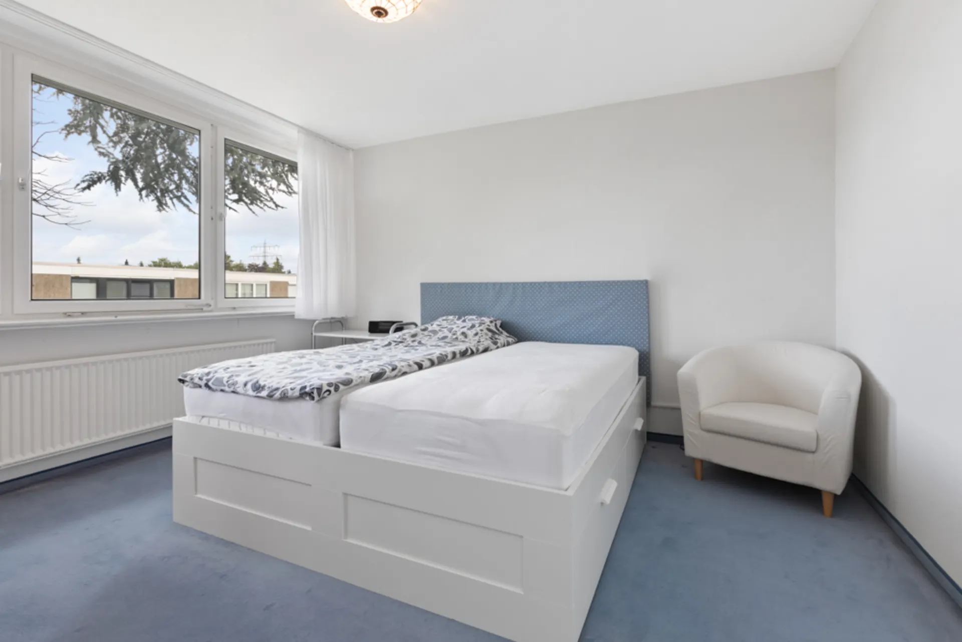 Helles Schlafzimmer mit großem Bett, blauem Teppich und weißem Sessel neben dem Fenster.
