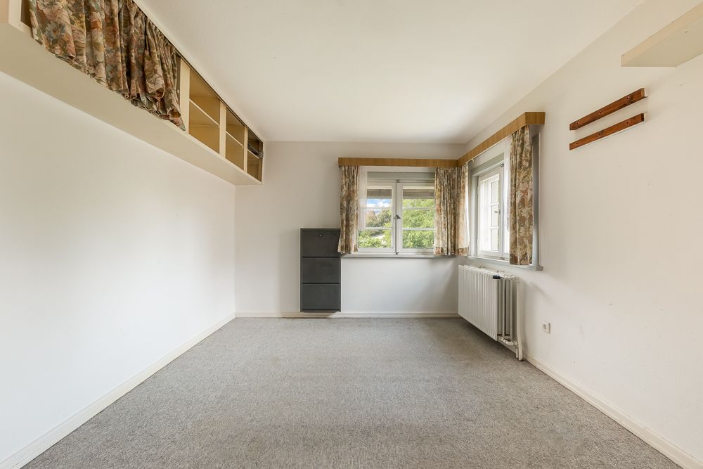 Leeres Zimmer mit Teppichboden, zwei Fenstern, Heizung, Vorhängen und offenen Wandregalen.