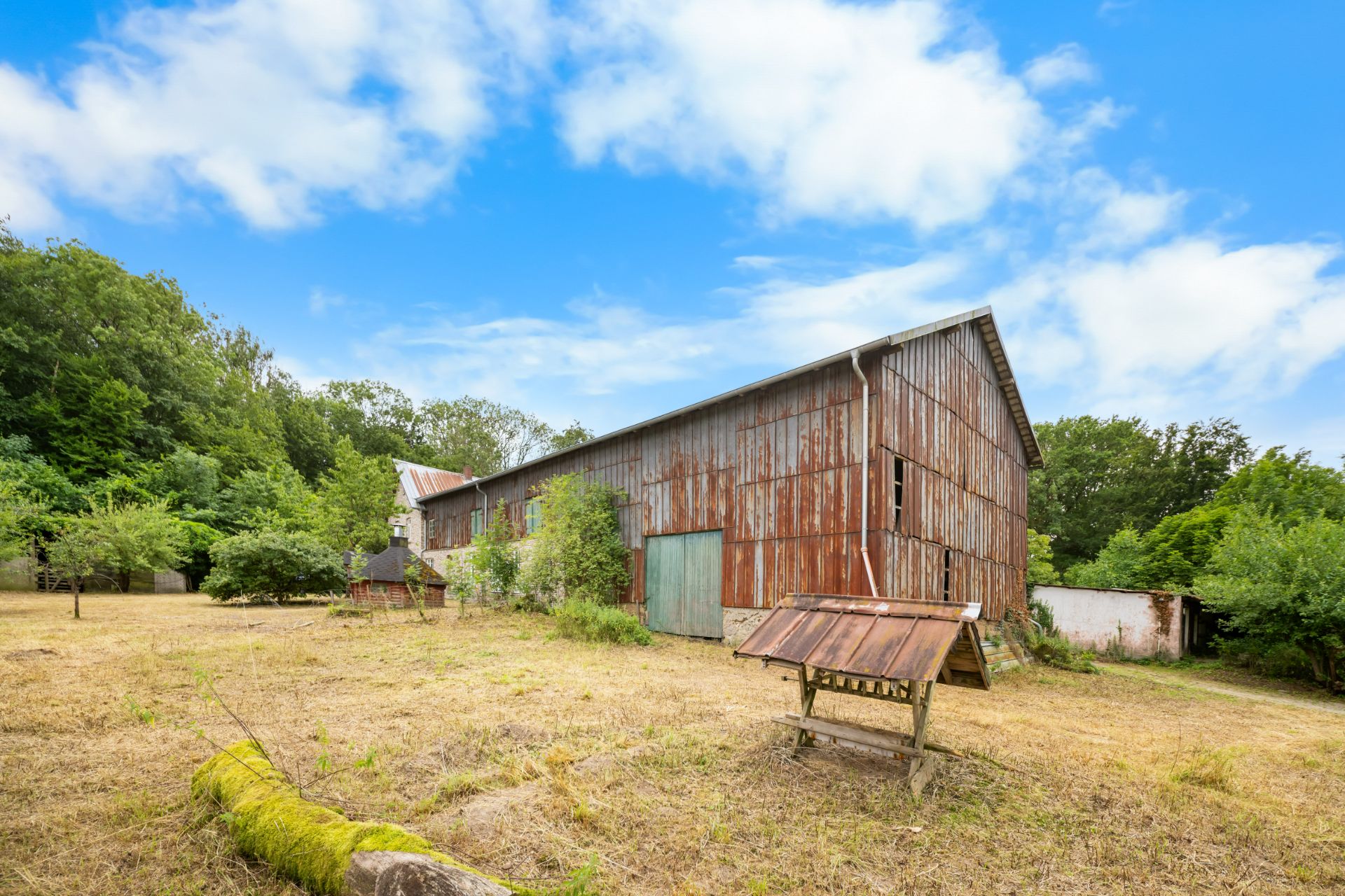 Große Scheune aus Holz mit rostiger Fassade und grünem Tor, umgeben von Bäumen auf trockenem Boden.